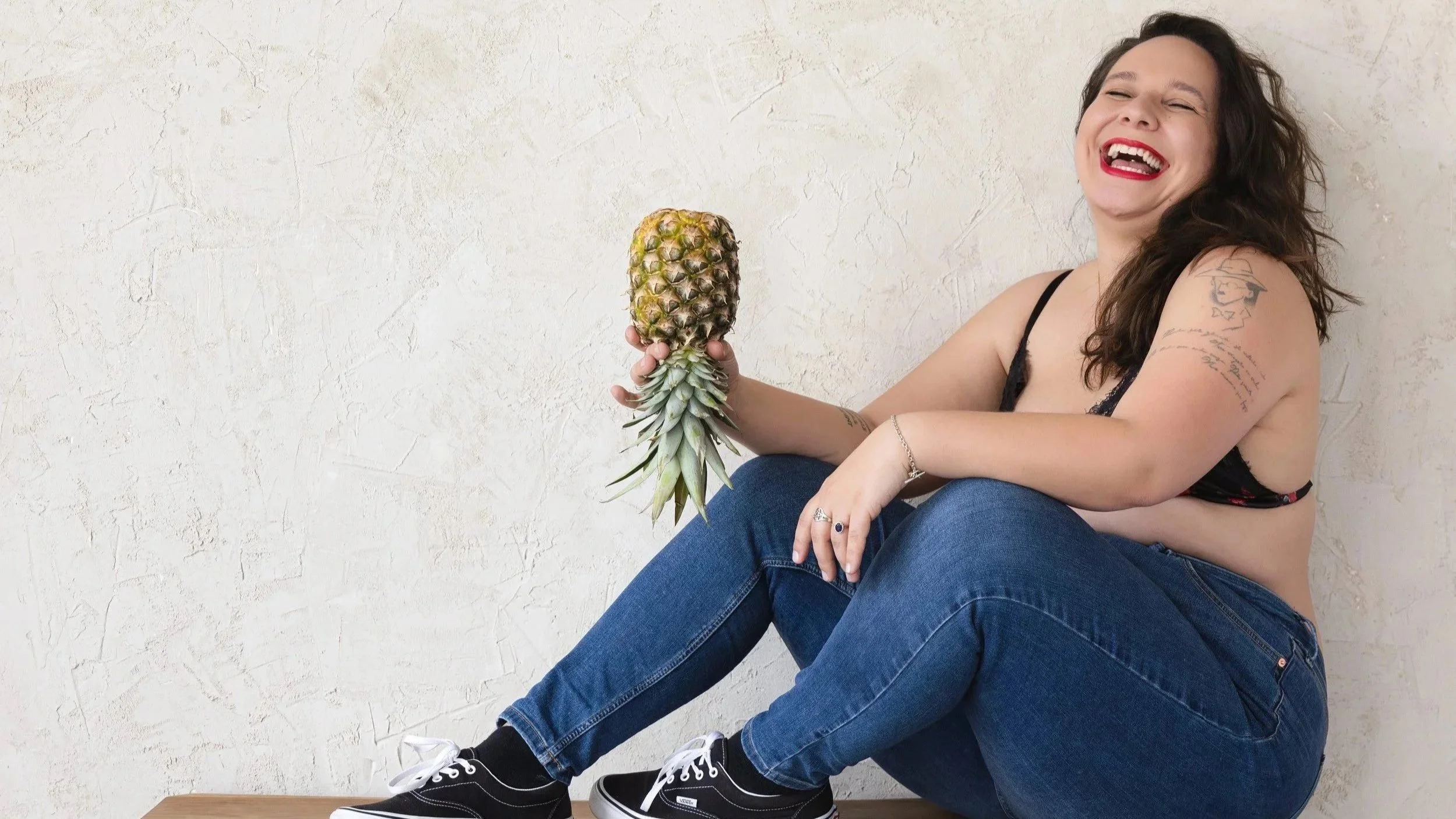 A woman with black hair, red lipstick, and tattoos on her arm holding a pineapple while sitting against a white textured wall, wearing a black top, blue jeans, and black sneakers.
