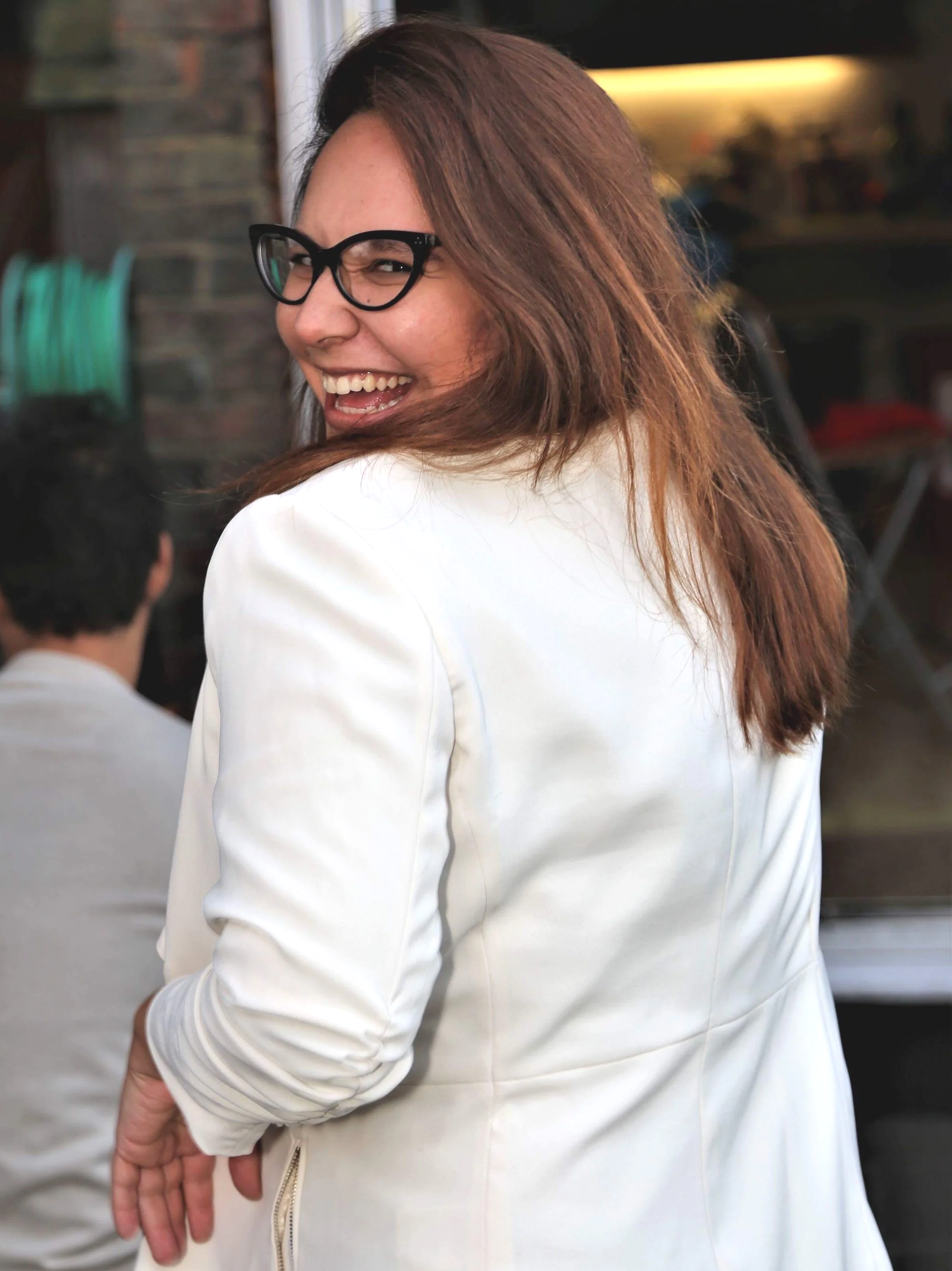 A woman with brown hair, black glasses, and a white jacket laughing and looking over her shoulder at the camera in a social setting.