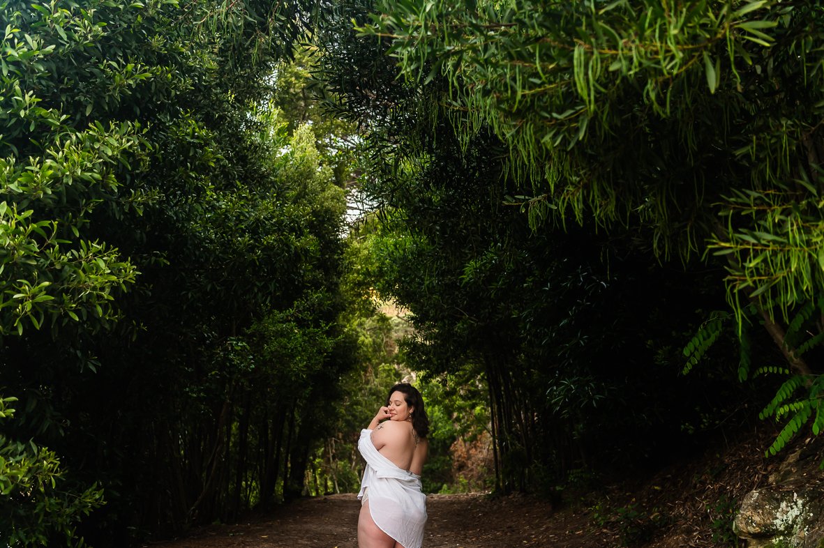 Woman doing a boudoir photoshoot standing on a forest trail, wrapped in a white towel, surrounded by dense green trees and foliage.