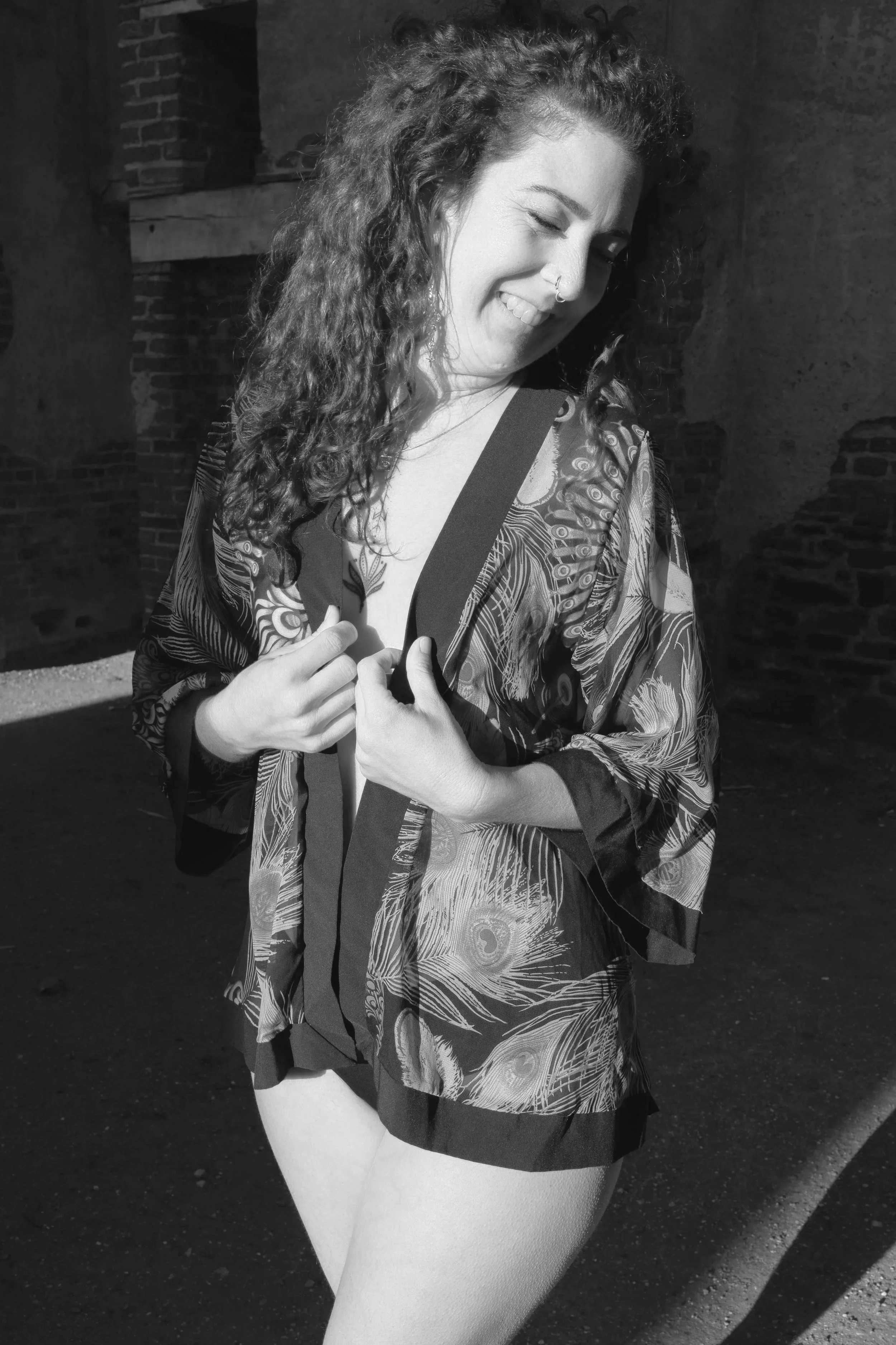 A woman with curly hair smiling and looking down, holding the edges of her kimono-style robe with peacock feather print, standing outdoors beside a brick building.