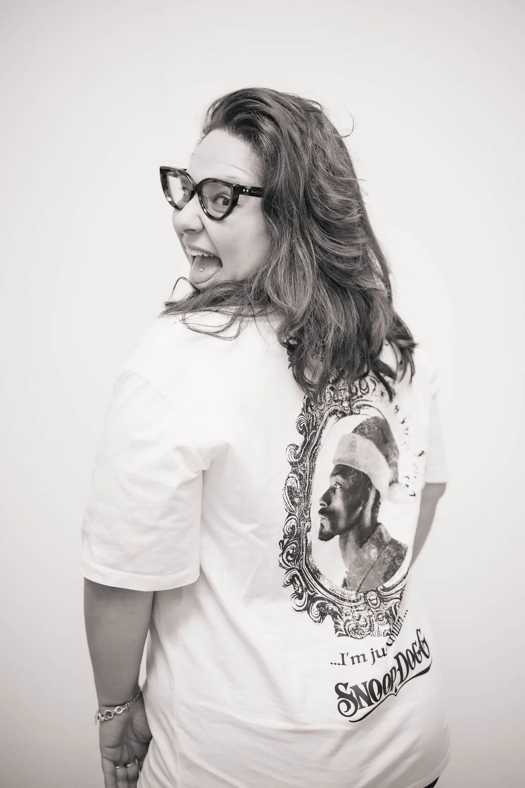 Black and white photo of a woman with glasses, winking and sticking out her tongue, wearing a Snoop Dog T-shirt with a portrait of a man in a hat on the back.