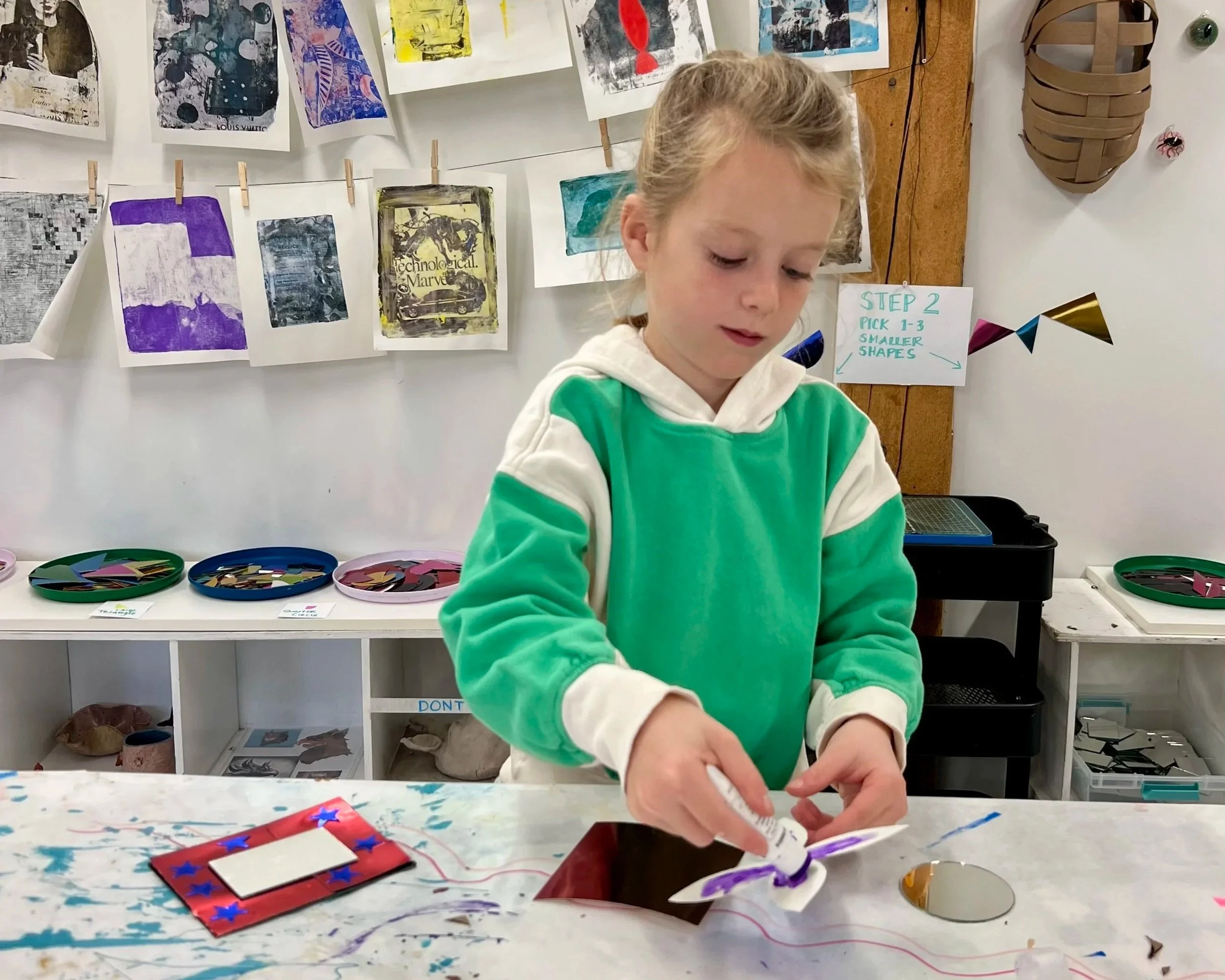 A young girl with blonde hair wearing a green and white hoodie is working on an art project at a table filled with supplies, in an art classroom with colorful artwork or prints hanging on the wall behind her.