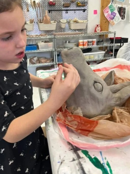 A young girl working on a dinosaur sculpture in an art studio, with shelves of art supplies in the background and the dinosaur wrapped in paper in front of her.