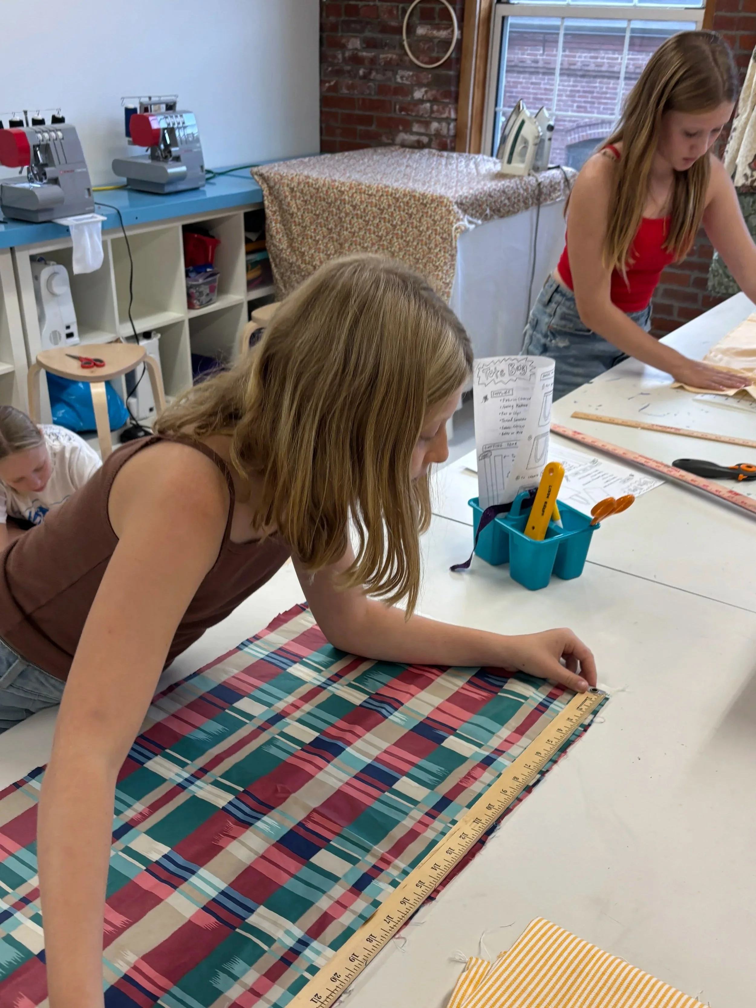 Two girls working on a sewing project with fabric and rulers, in a crafting room.