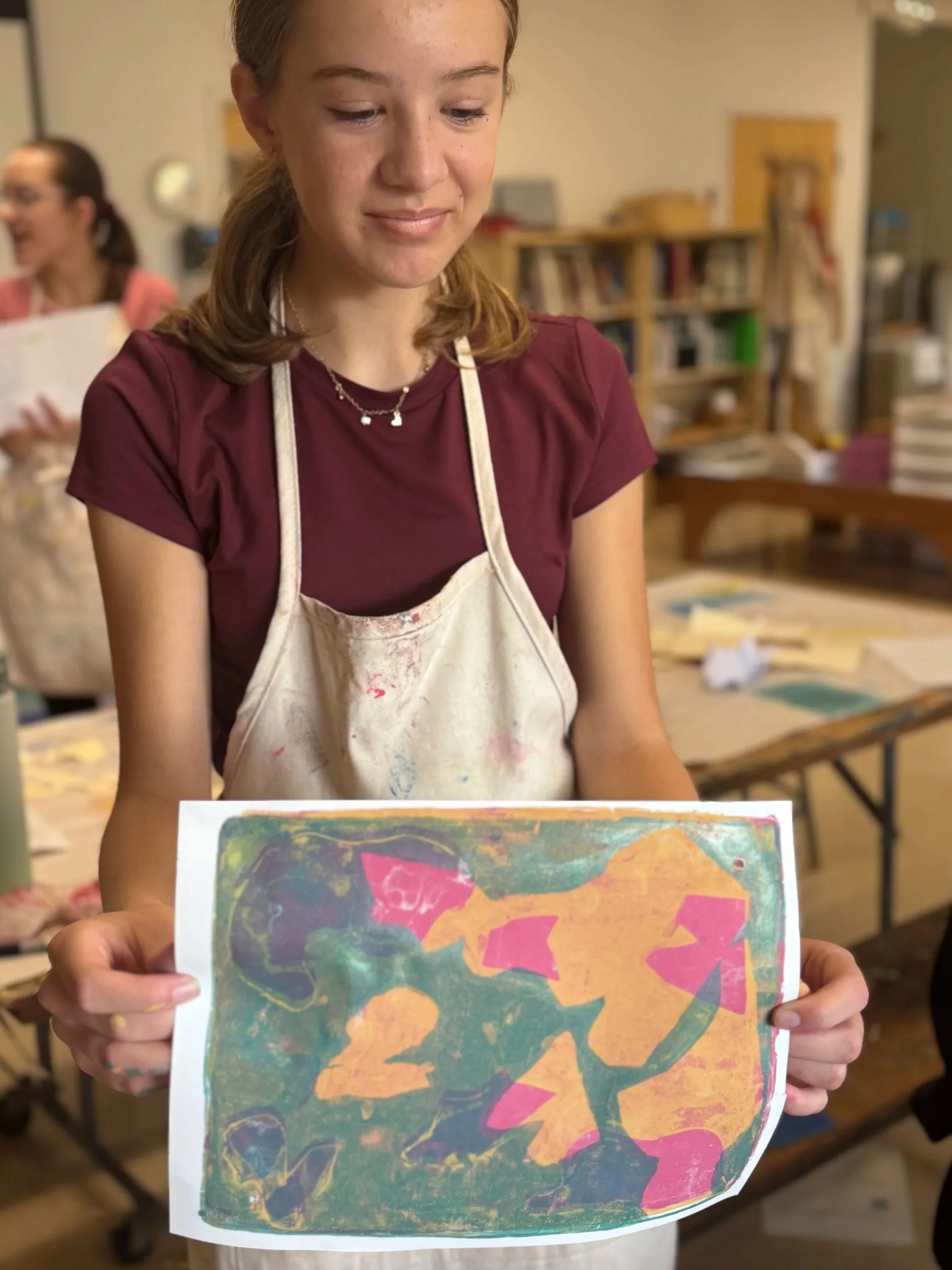 Young girl in a maroon shirt and apron holding a colorful abstract artwork on paper in an art classroom.