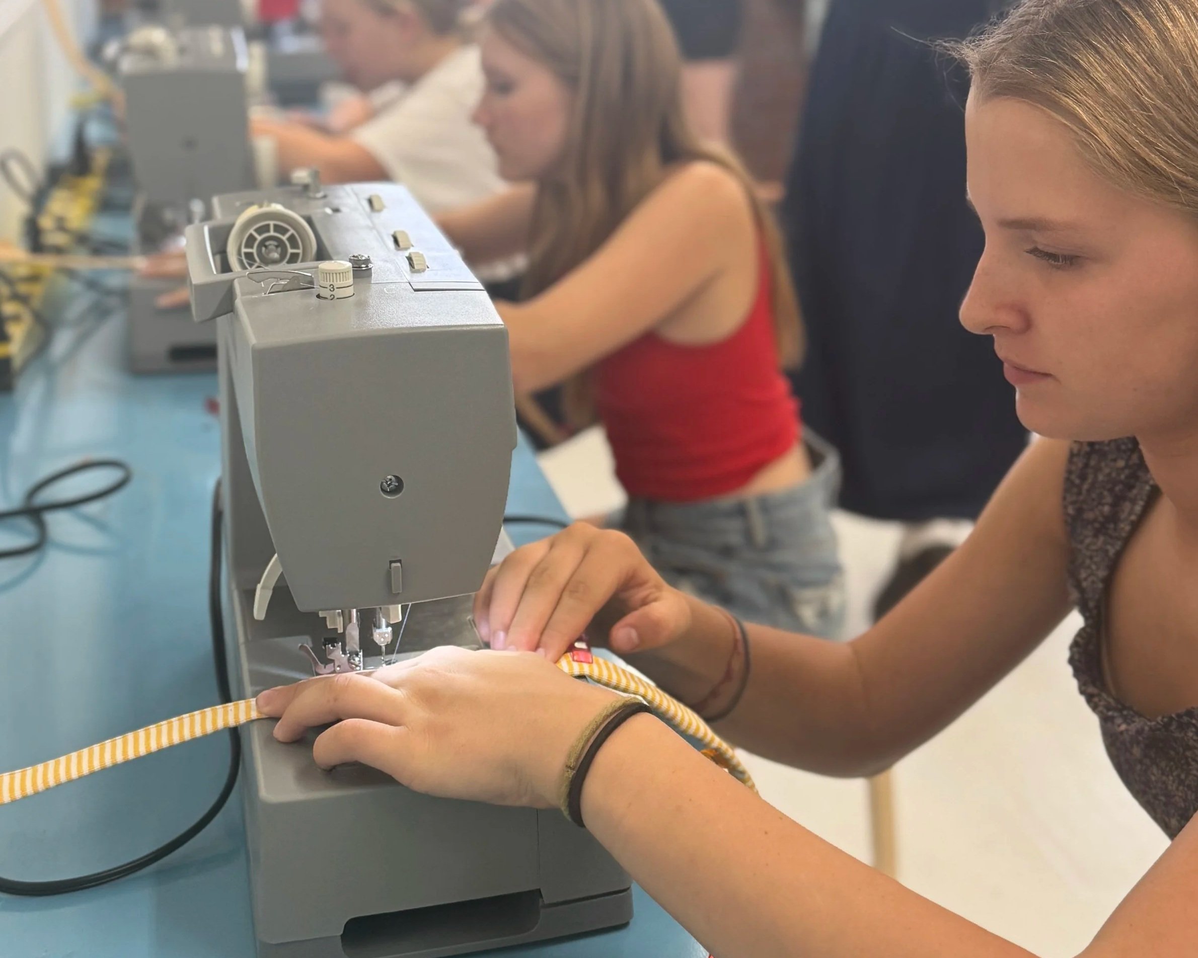 Teen girl sewing a yellow and white striped fabric on a sewing machine in a classroom with other students working on sewing projects.