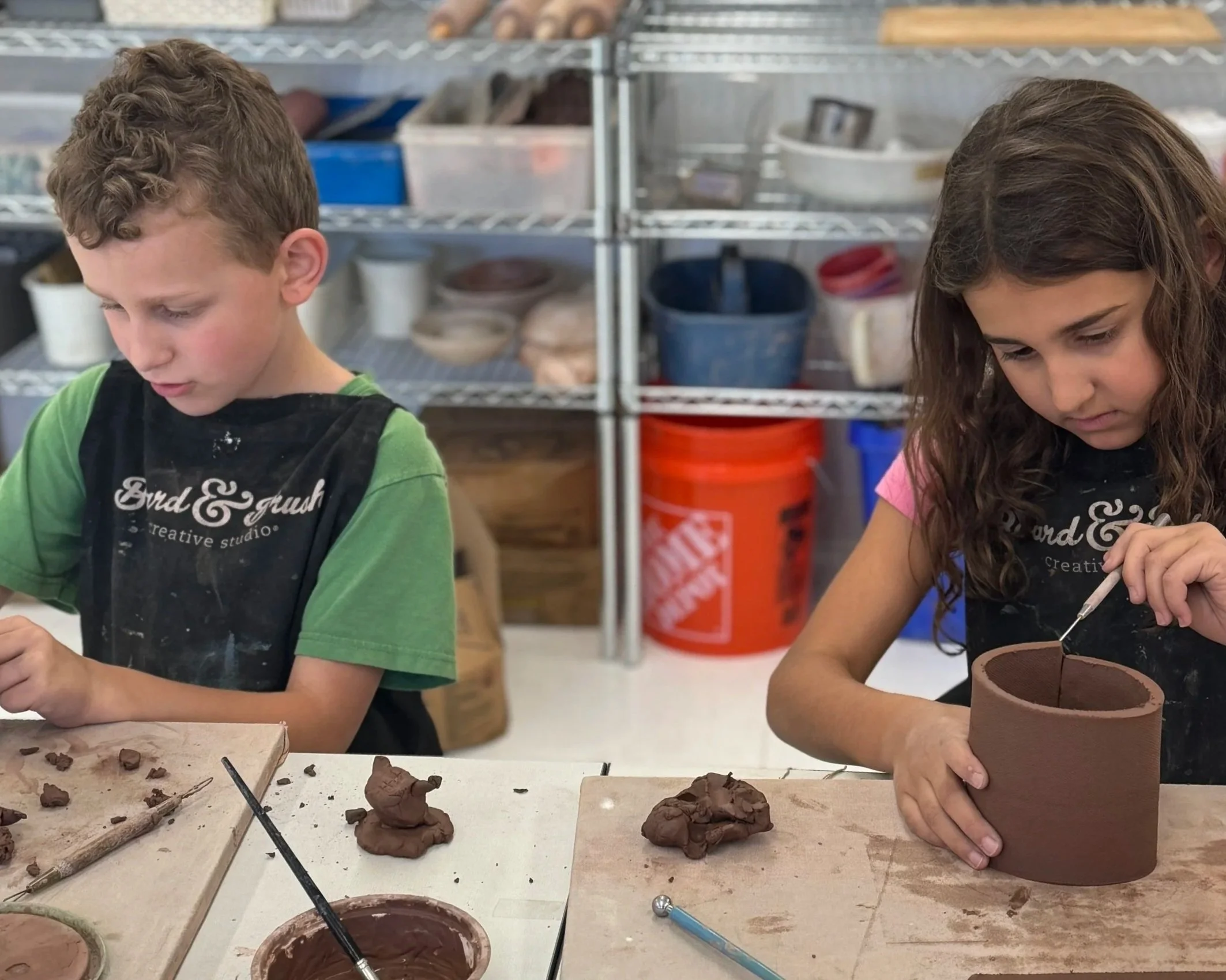 Two children working on clay pottery projects at a workshop table. The boy on the left has curly hair and is focused on sculpting, while the girl on the right has long, wavy hair and is shaping a cylindrical clay piece. There are various clay tools a