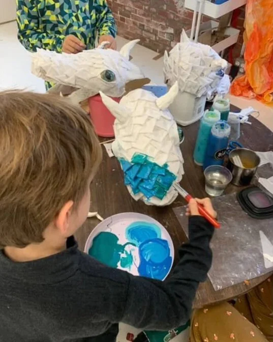 Child painting a paper-mâché unicorn sculpture with blue paint at an art table.