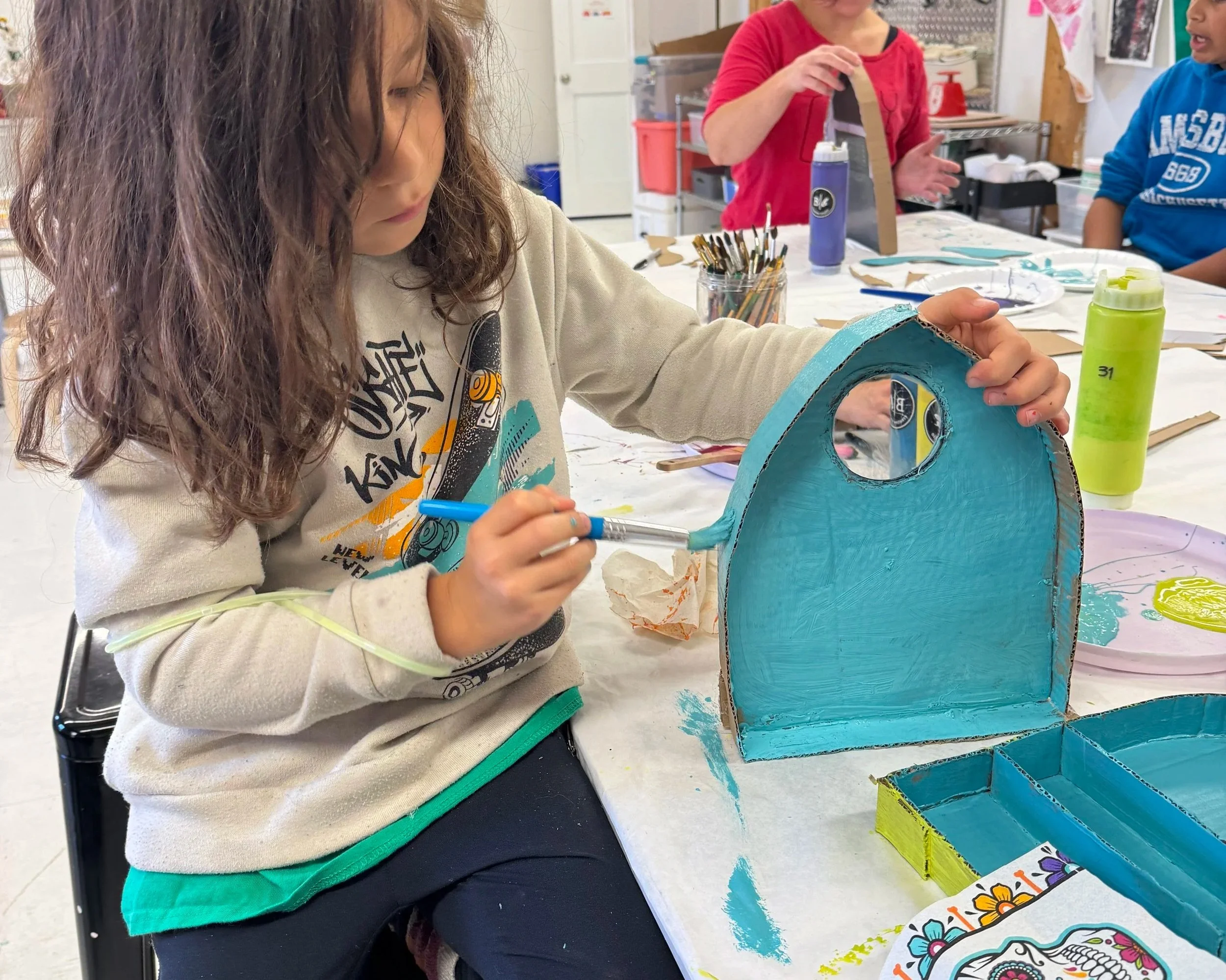 A young girl painting a cardboard pirate skull mask blue at a craft table, with other children and art supplies in the background.