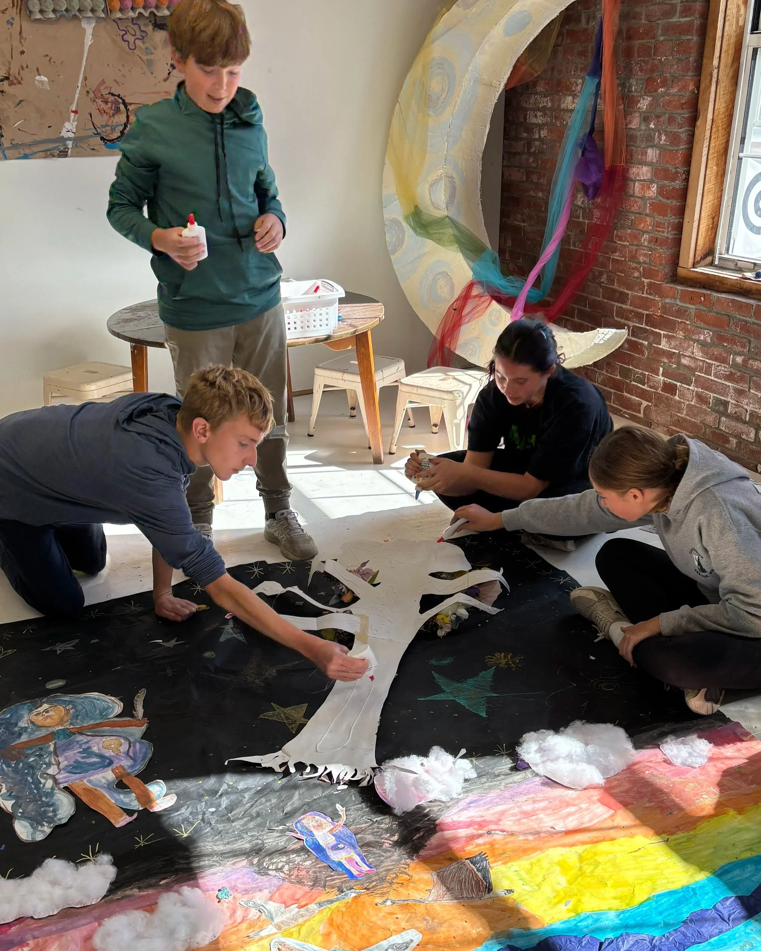 Group of five people working on an art project on a large black paper with colorful drawings and cutouts, during daytime in a room with brick and white walls, a circular opening with colorful fabric, and sunlight coming through a window.