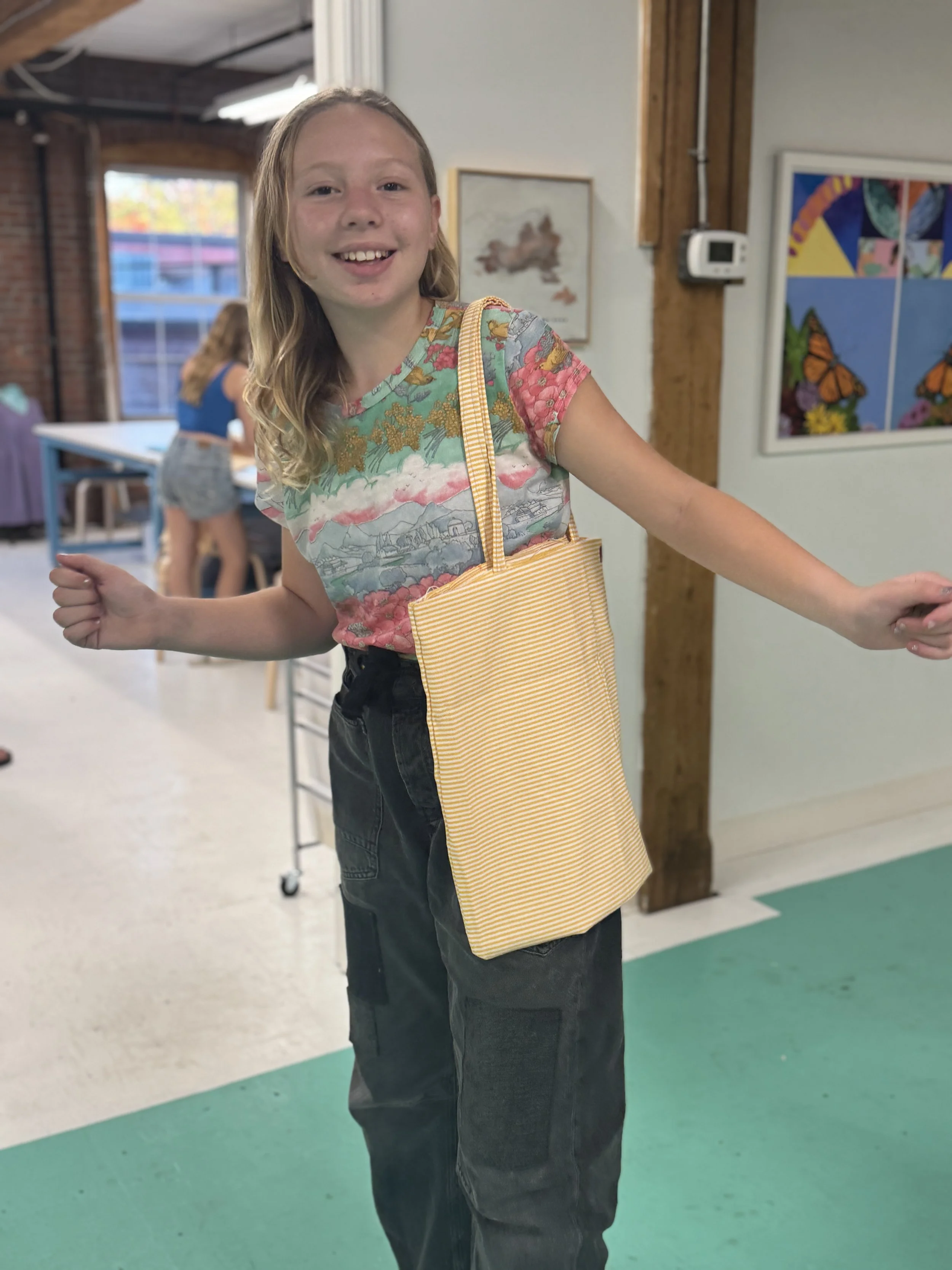 Smiling girl with long blonde hair wearing a colorful top and black pants, carrying a yellow striped tote bag, inside a room with artwork on the walls.