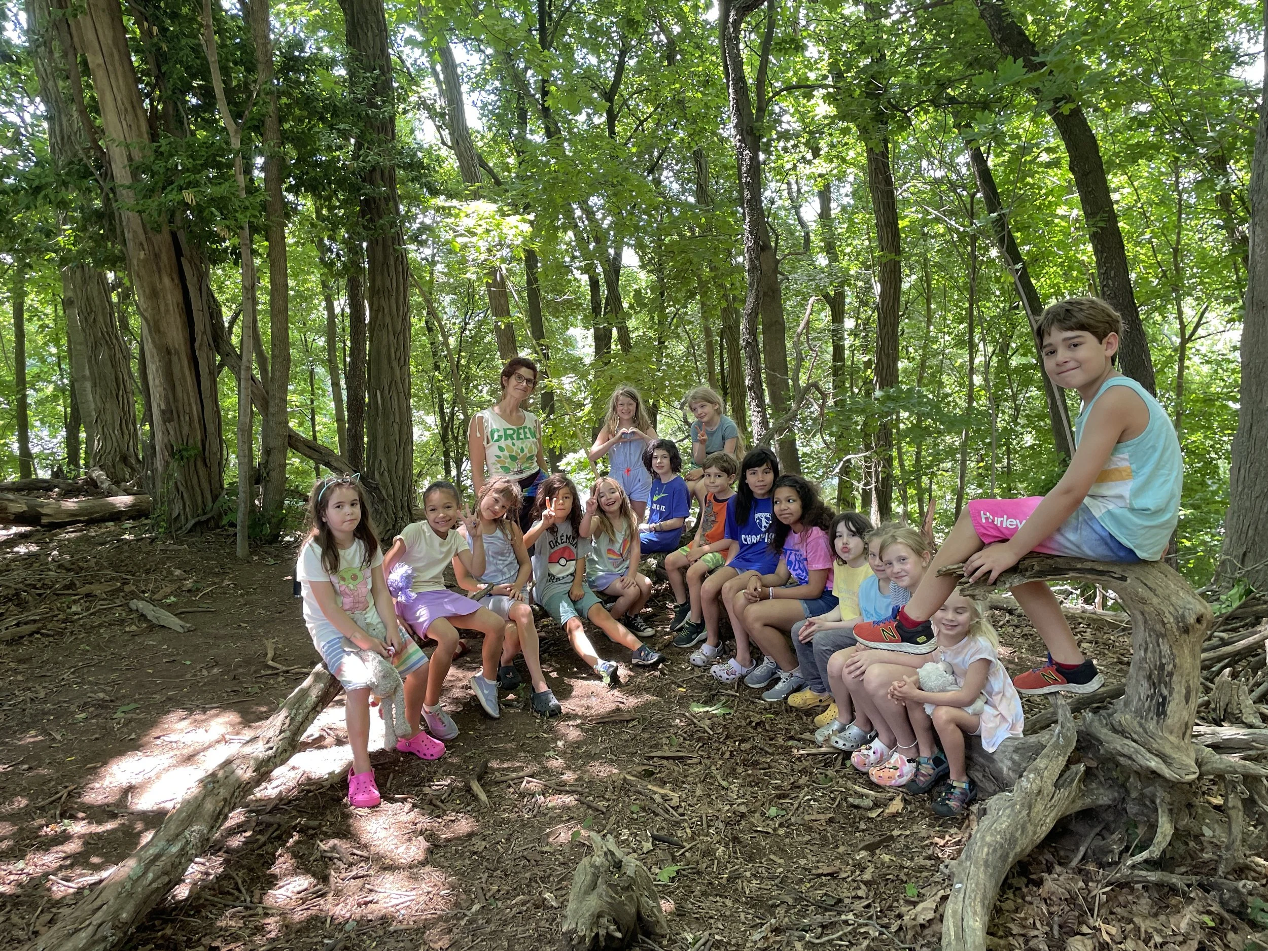 Group of children and a woman sitting on fallen tree branches in a dense green forest, smiling and posing for the photo.