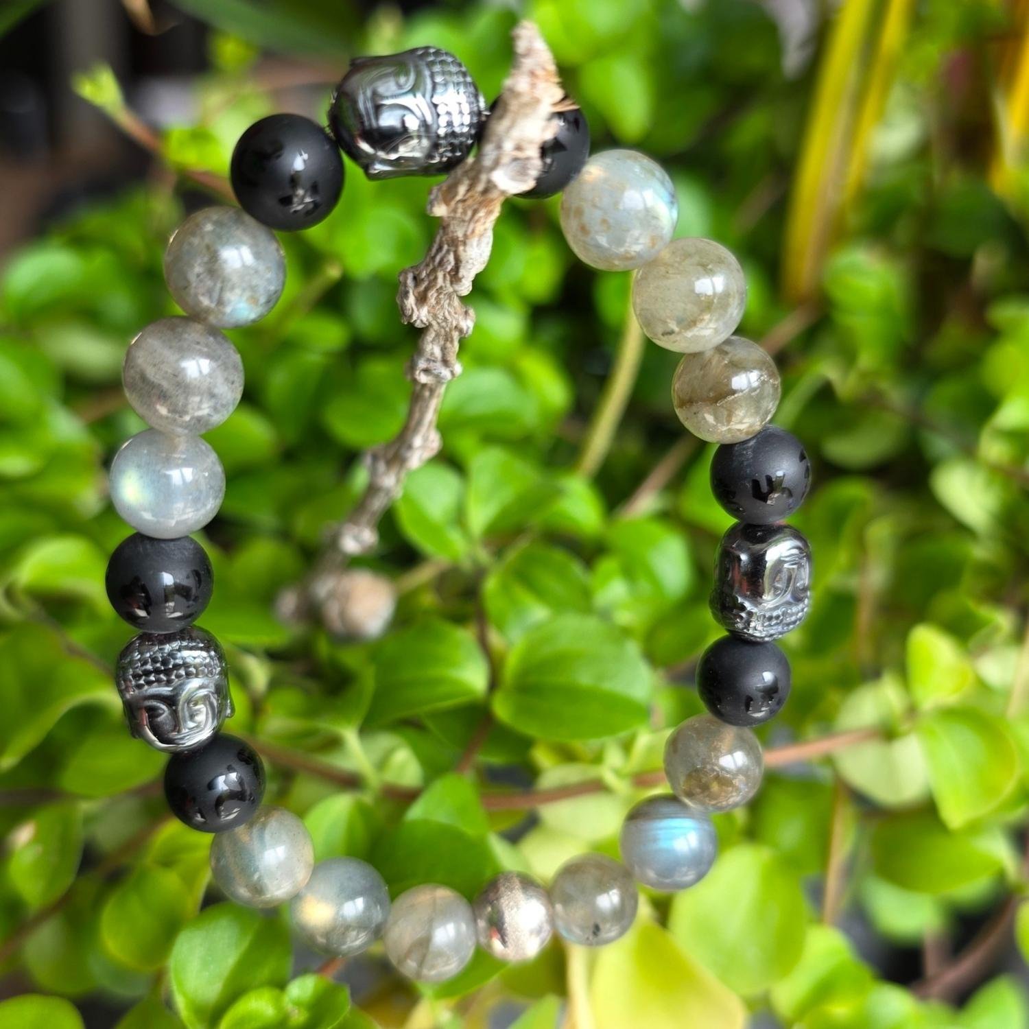 Buddha Mantra Armband aus Labradorit, Onyx mit tibetischen Zeichen und Hämatit Buddha-Köpfen, hängend vor grünen Pflanzen
