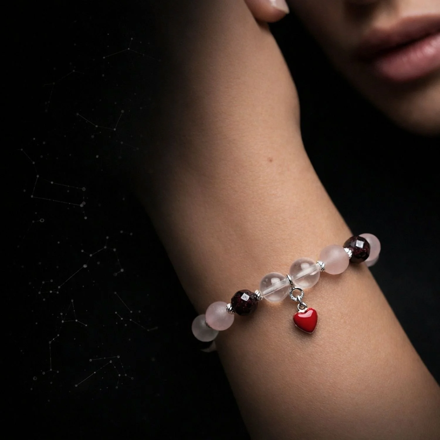 Sternenarmband aus mattem Rosenquarz, facettiertem Granat und klarem Bergkristall mit 925 Silber Zwischenelementen und rotem Herzanhänger, getragen am Handgelenk einer Frau in hochwertiger Studioaufnahme