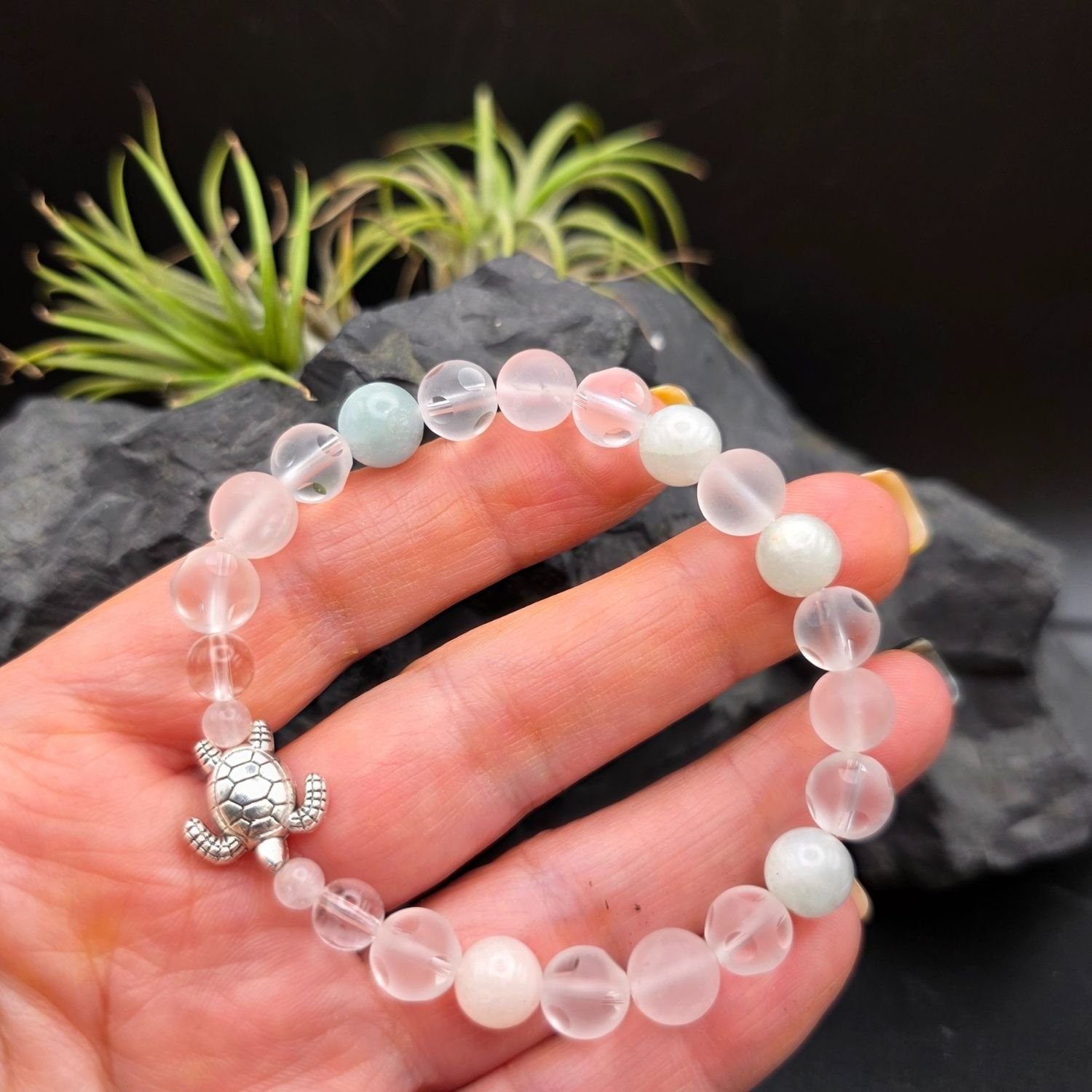 Schildkröten Armband in einer Hand gehalten, zeigt mattierten Bergkristall, Aquamarin, Mondstein, Rosenquarz und den Schildkröten-Anhänger