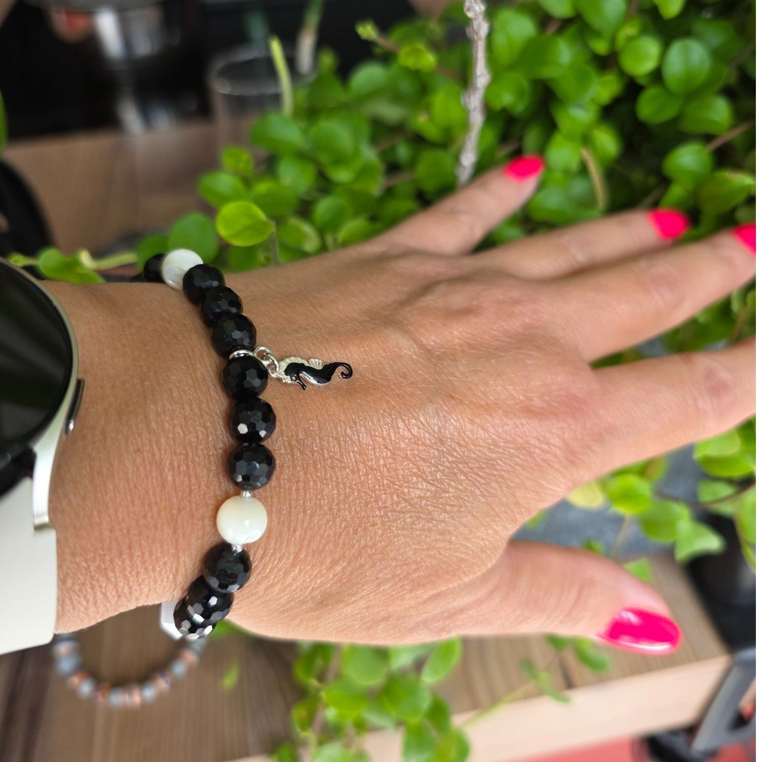 Elegantes Armband aus schwarzem Turmalin und weißen Perlen mit emailliertem Seepferdchen-Anhänger an einem Handgelenk mit Uhr.