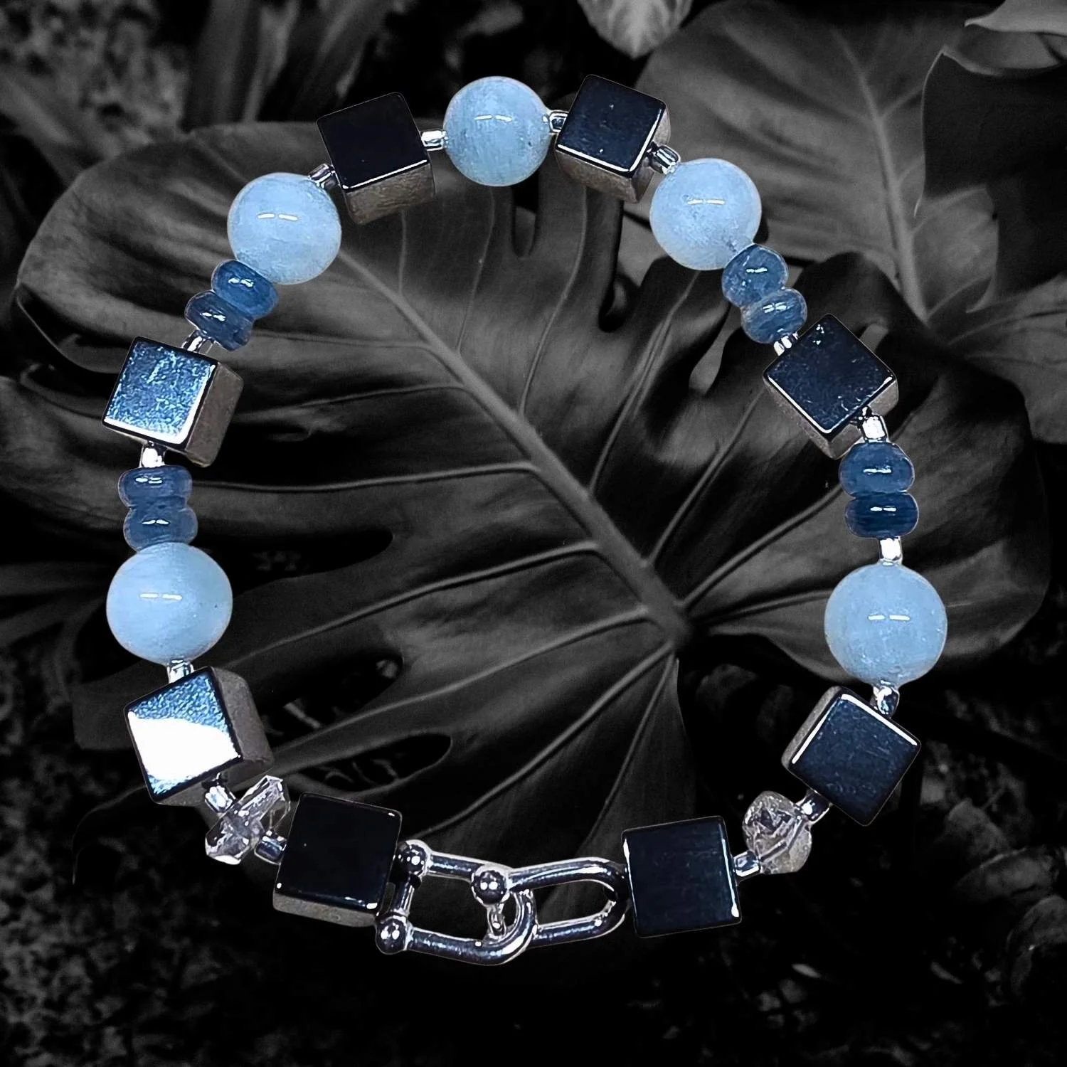 Herrenarmband Nautische Alchemy aus Hämatit, Aquamarin, Kyanit und Herkimer Quarz mit 925 Silber Bootsschäkel auf dunklem Blatt arrangiert