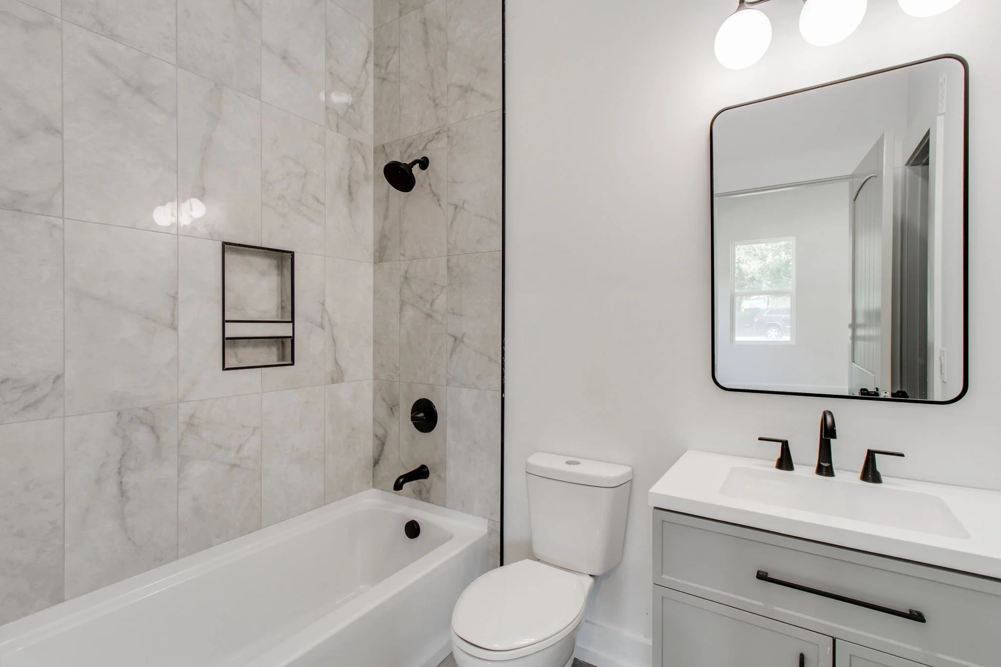 Modern bathroom with bathtub, gray tiled shower wall, white toilet, white vanity with black fixtures, and rectangular mirror.