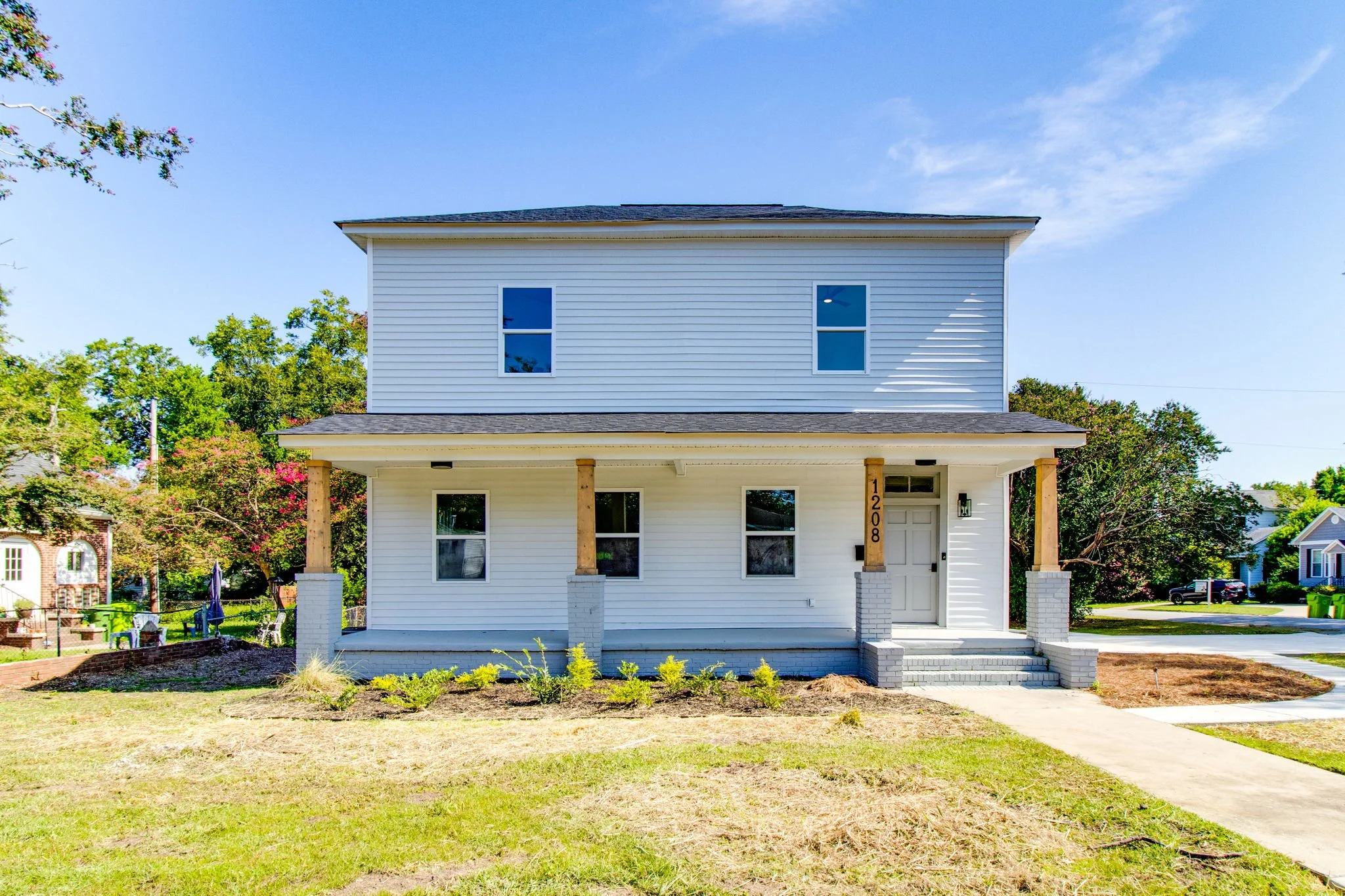 Two-story white house with front porch and number 1208 on column, surrounded by green lawn and trees.