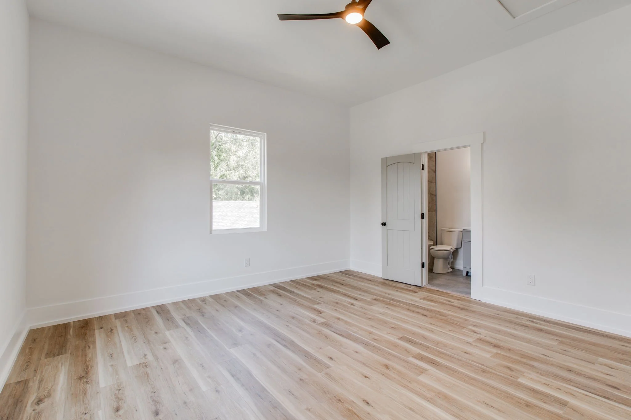 Empty room with wooden flooring, white walls, a window, ceiling fan, and an open door leading to a bathroom.