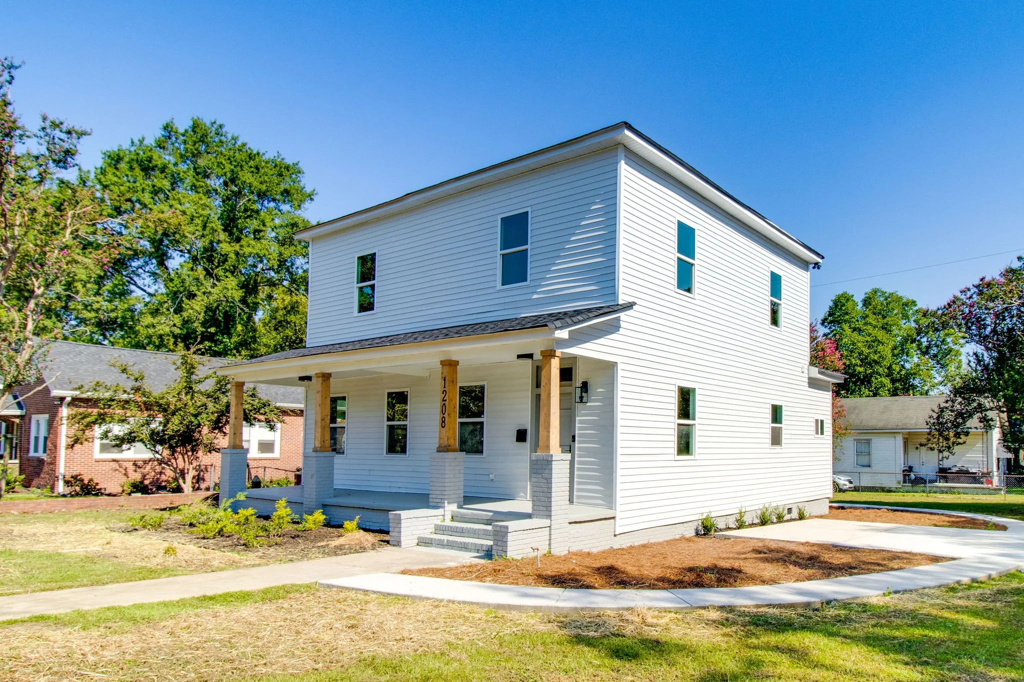 Newly constructed white two-story house with wood columns and a small front porch, surrounded by a lawn, with neighboring houses and trees in the background.