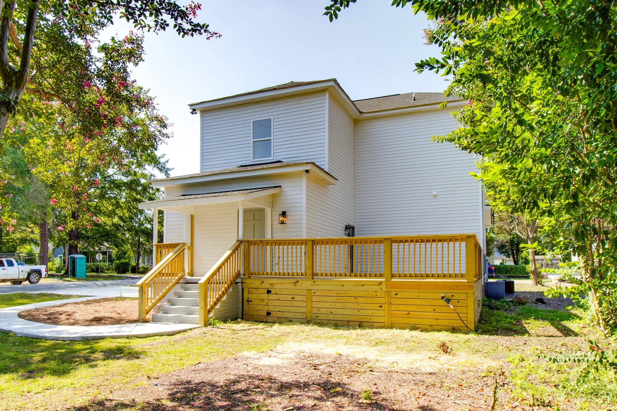 White house with wooden porch and railing, surrounded by trees and greenery, with an outdoor portable toilet and pickup truck visible in the background.