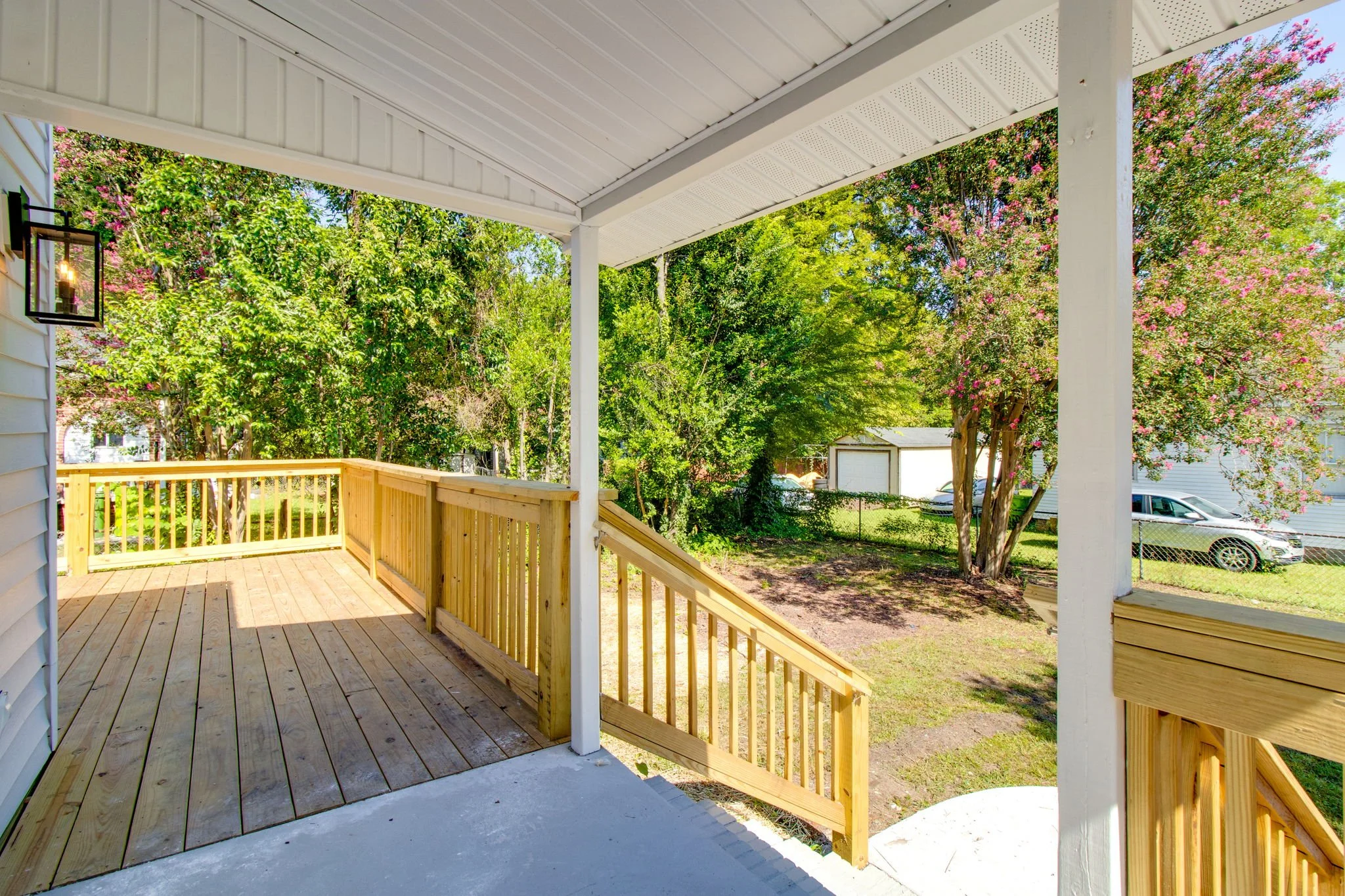 Wooden porch with railing, overlooking a garden area with trees and a vehicle in the background.