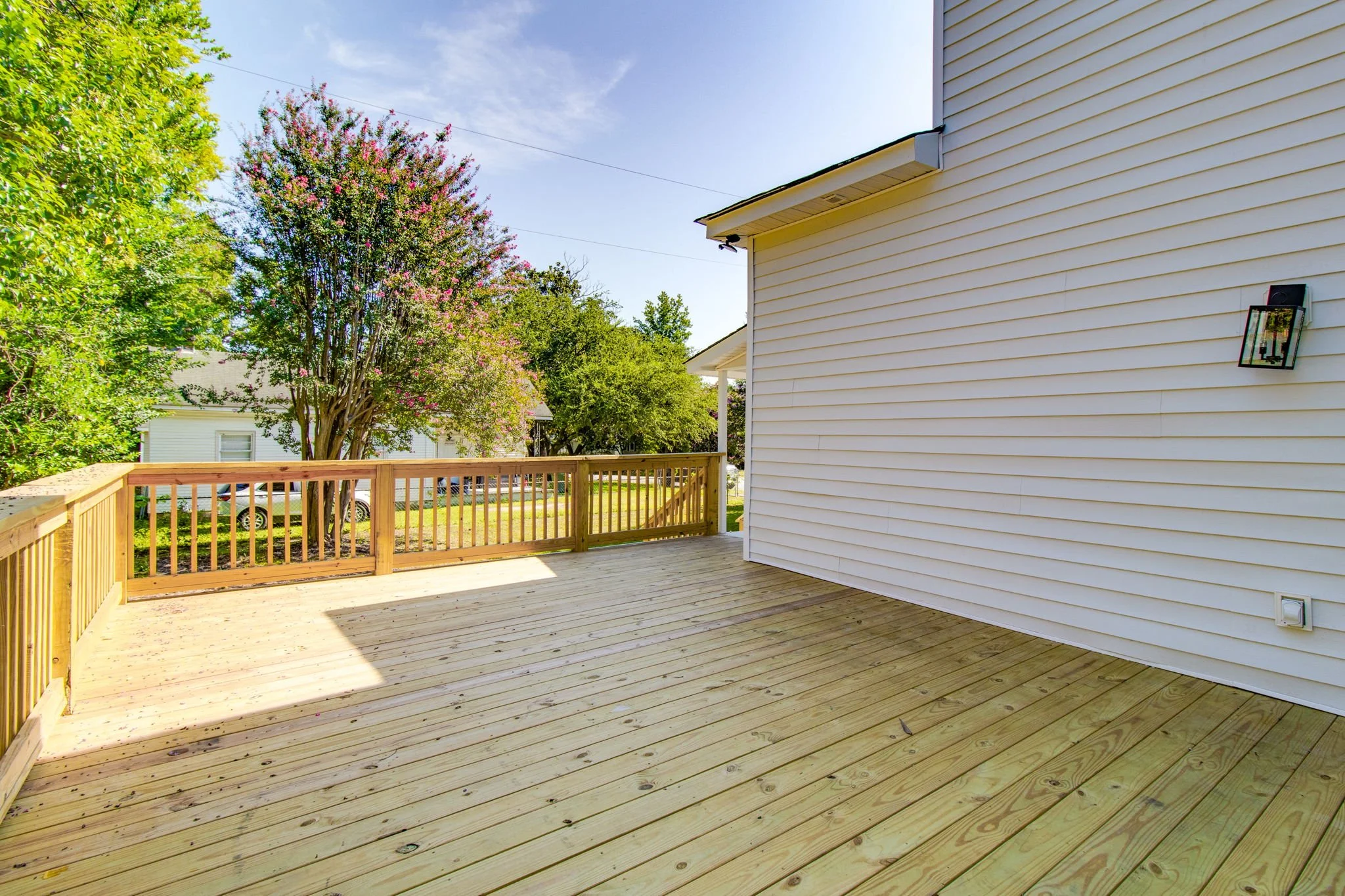 Wooden deck with railing attached to a white house, surrounded by green trees and a clear blue sky.