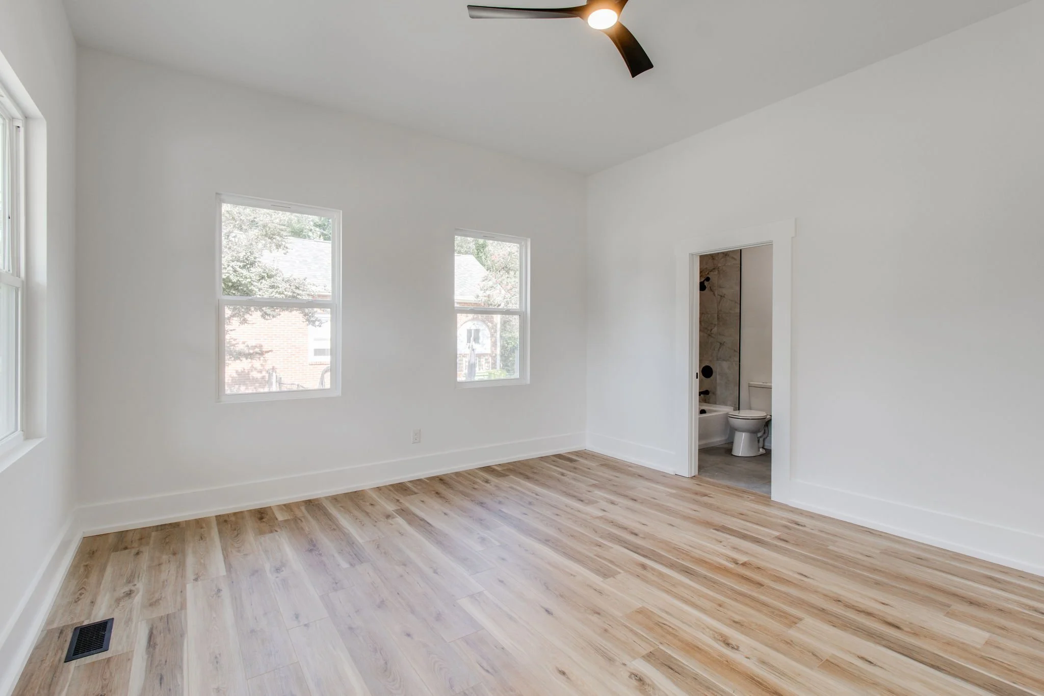 Empty room with hardwood floor, white walls, ceiling fan, two windows, and an open door to a bathroom with visible toilet.