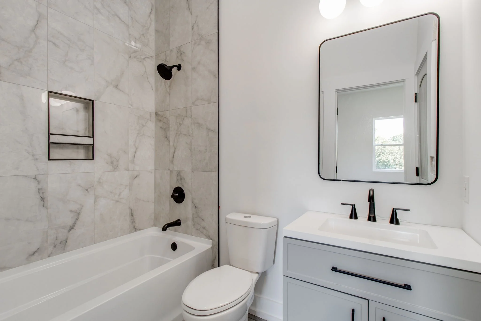 Modern bathroom with white bathtub, ceramic tiles, toilet, sink, rectangular mirror, black fixtures, and dark vanity handle.