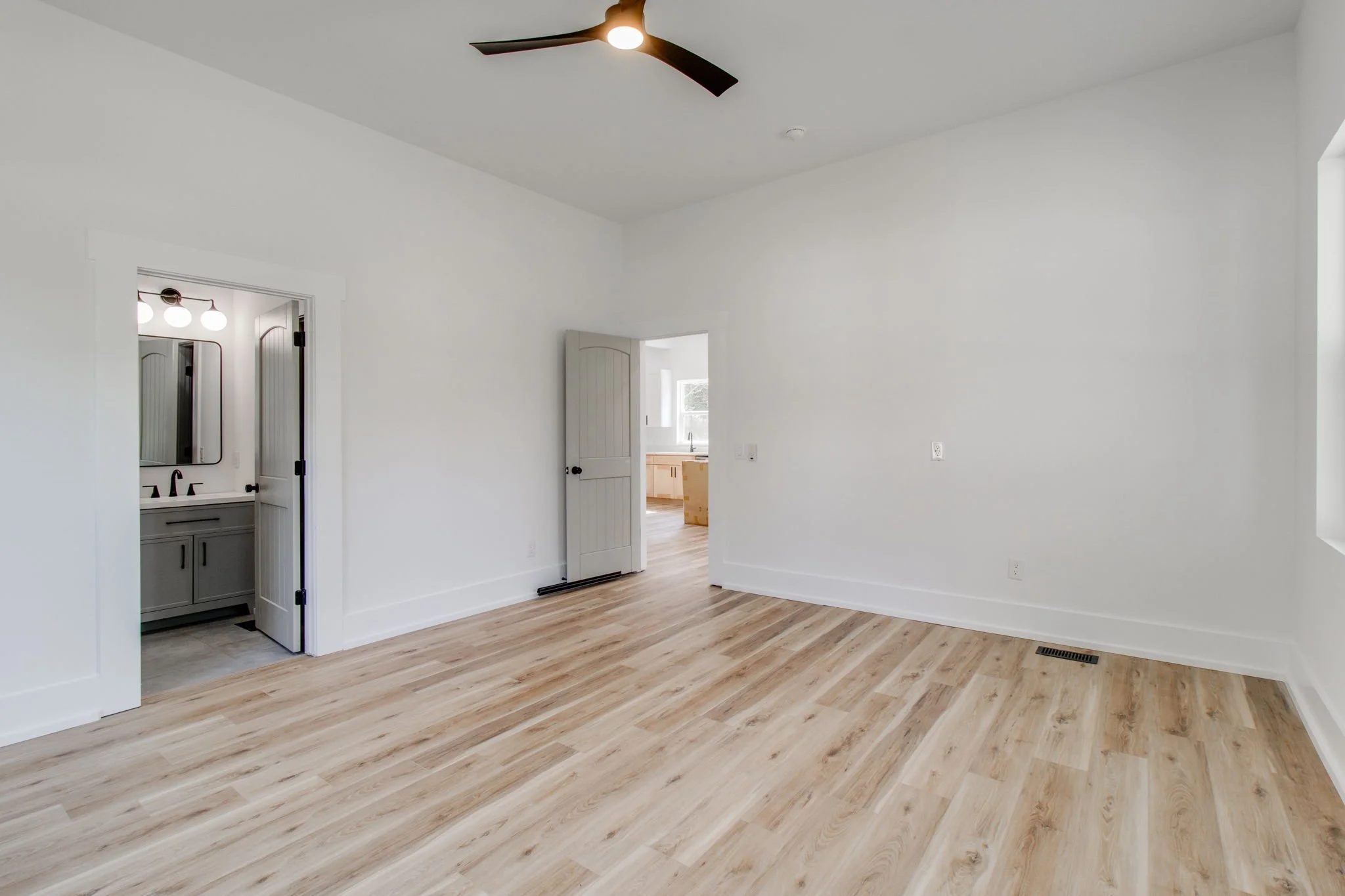 empty room with wood flooring, white walls, ceiling fan, open doors to bathroom and another room