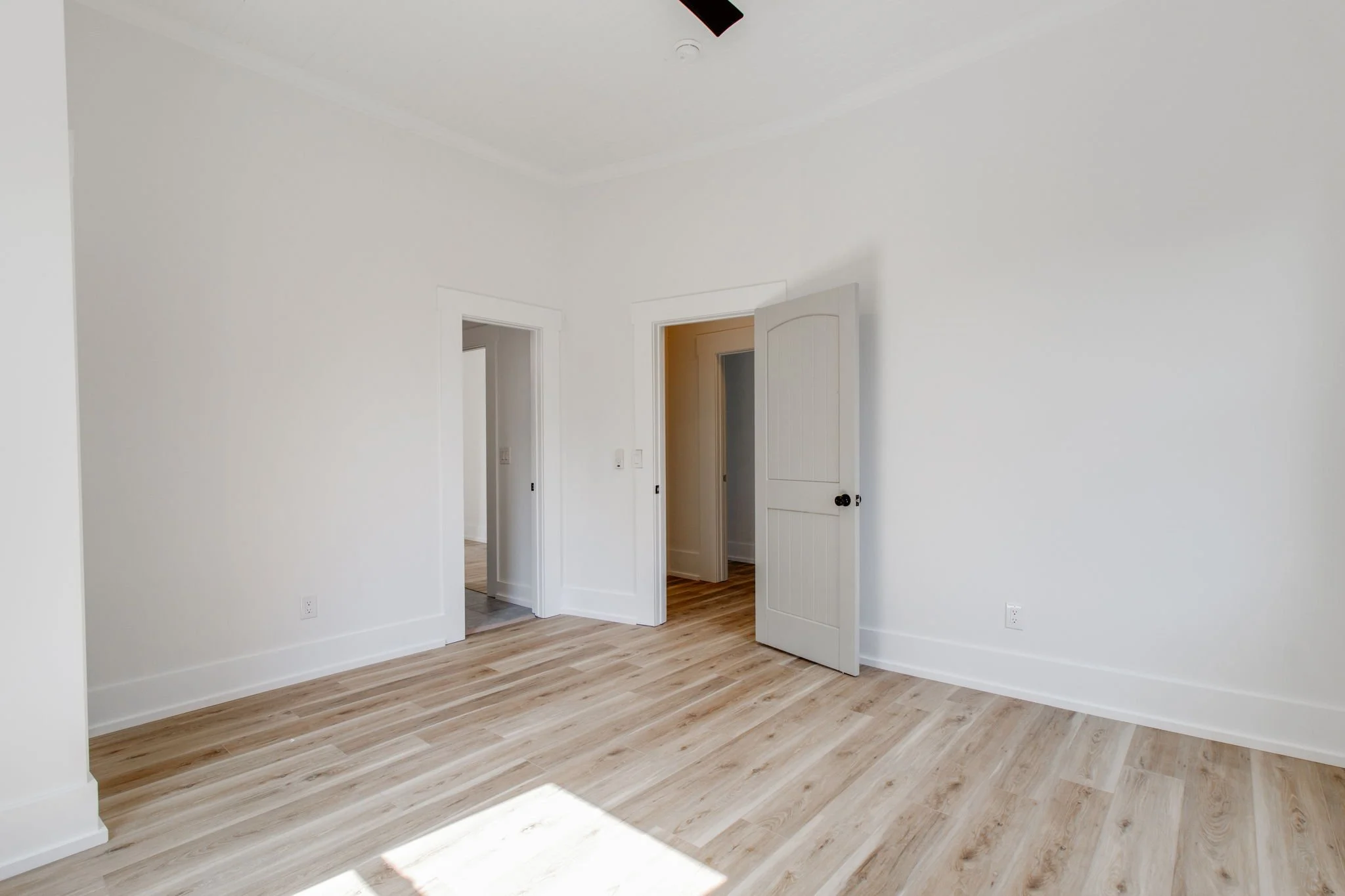 Minimalist empty room with light wood flooring, white walls, and two doorways.