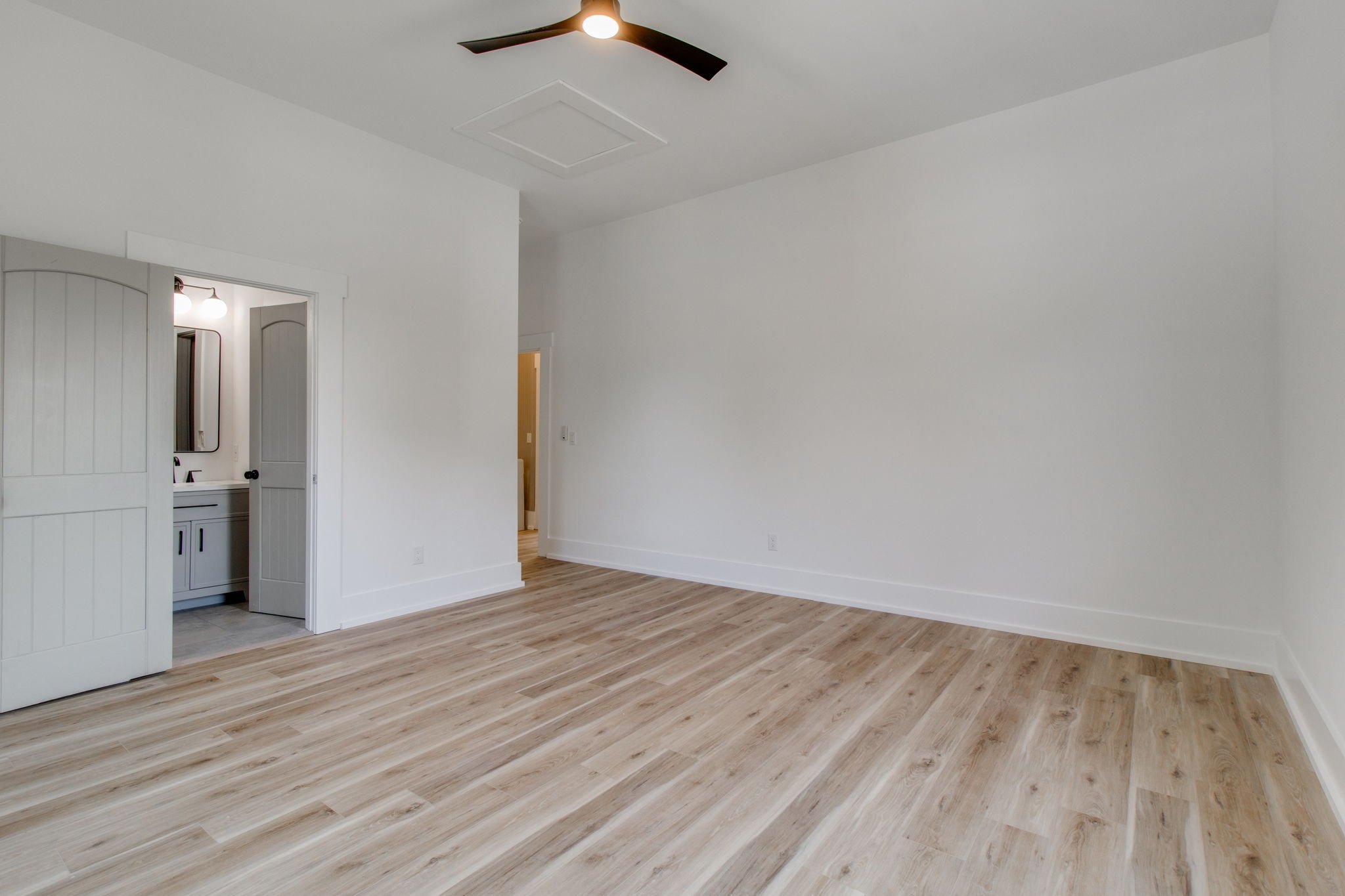 Empty room with light wooden flooring, white walls, ceiling fan, and an open door leading to a bathroom.