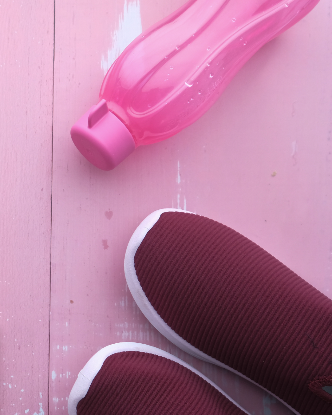 Pink water bottle and maroon sneakers with white soles on a pink wooden floor.