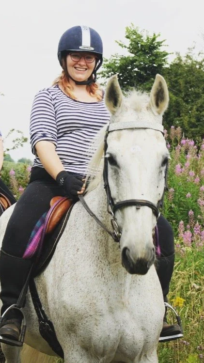 Woman riding a white horse, wearing a striped shirt, helmet, and glasses, in a garden with pink flowers and green trees.