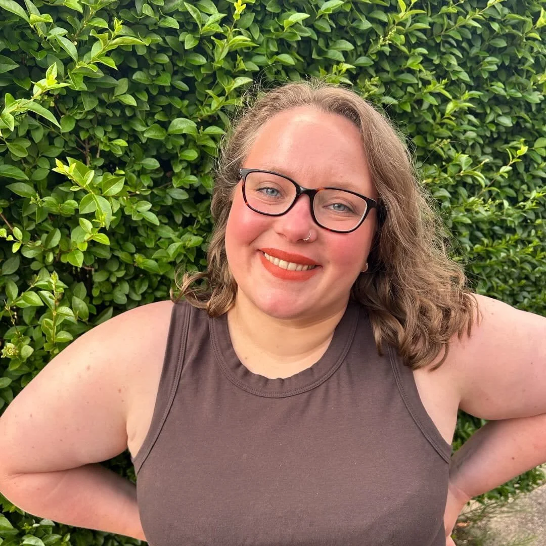 Claire Bonnet is a white female personal trainer who is wearing glasses has a nose piercing and wavy brown hair. She is smiling in front of a hedge.