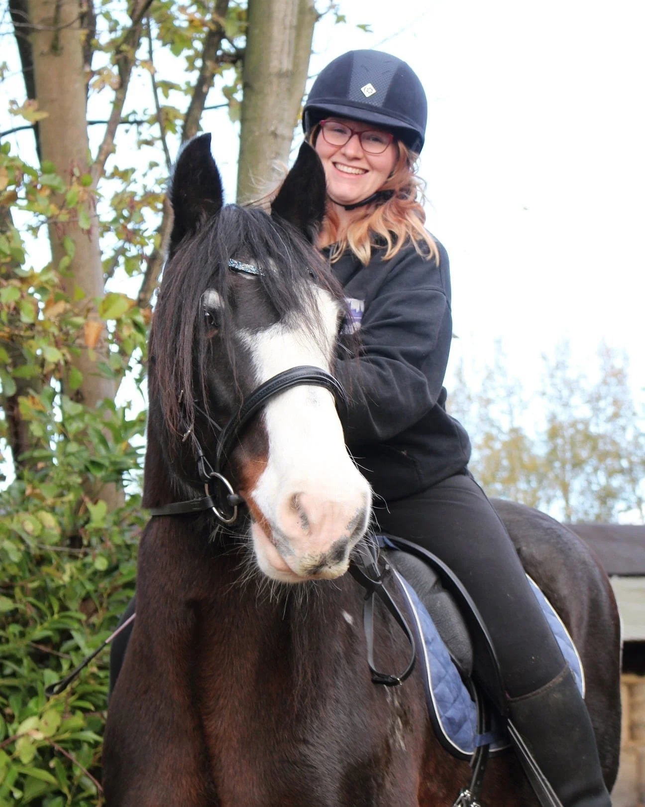 A young woman wearing a black helmet and glasses is riding a dark brown horse with a white face marking. She is smiling and outdoors near trees with green leaves.