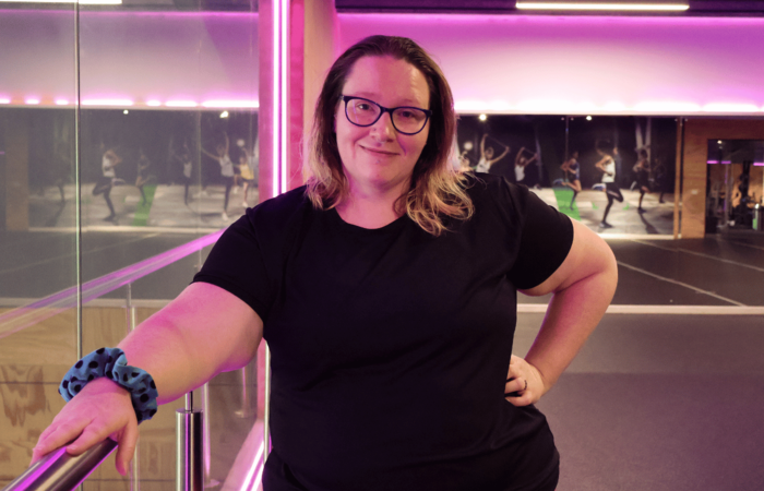 Rhiannon is a woman with glasses and shoulder-length hair smiling at the camera, wearing a black t-shirt, standing in a gym with pink and purple lighting, mirrored walls, and fitness equipment.