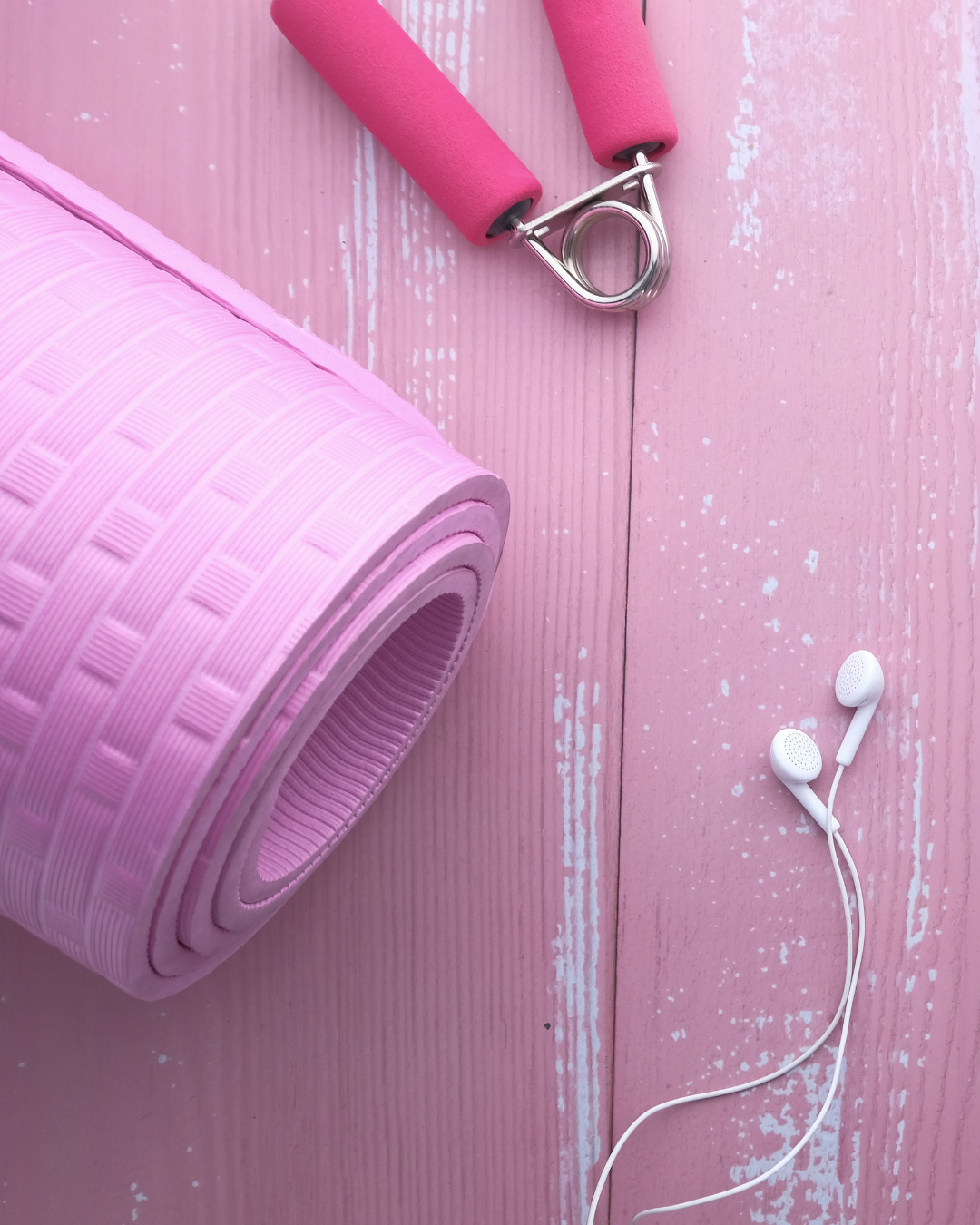 A pink yoga mat rolled up, a pink hand strengthener, and white earbud headphones are arranged on a pink wooden surface.