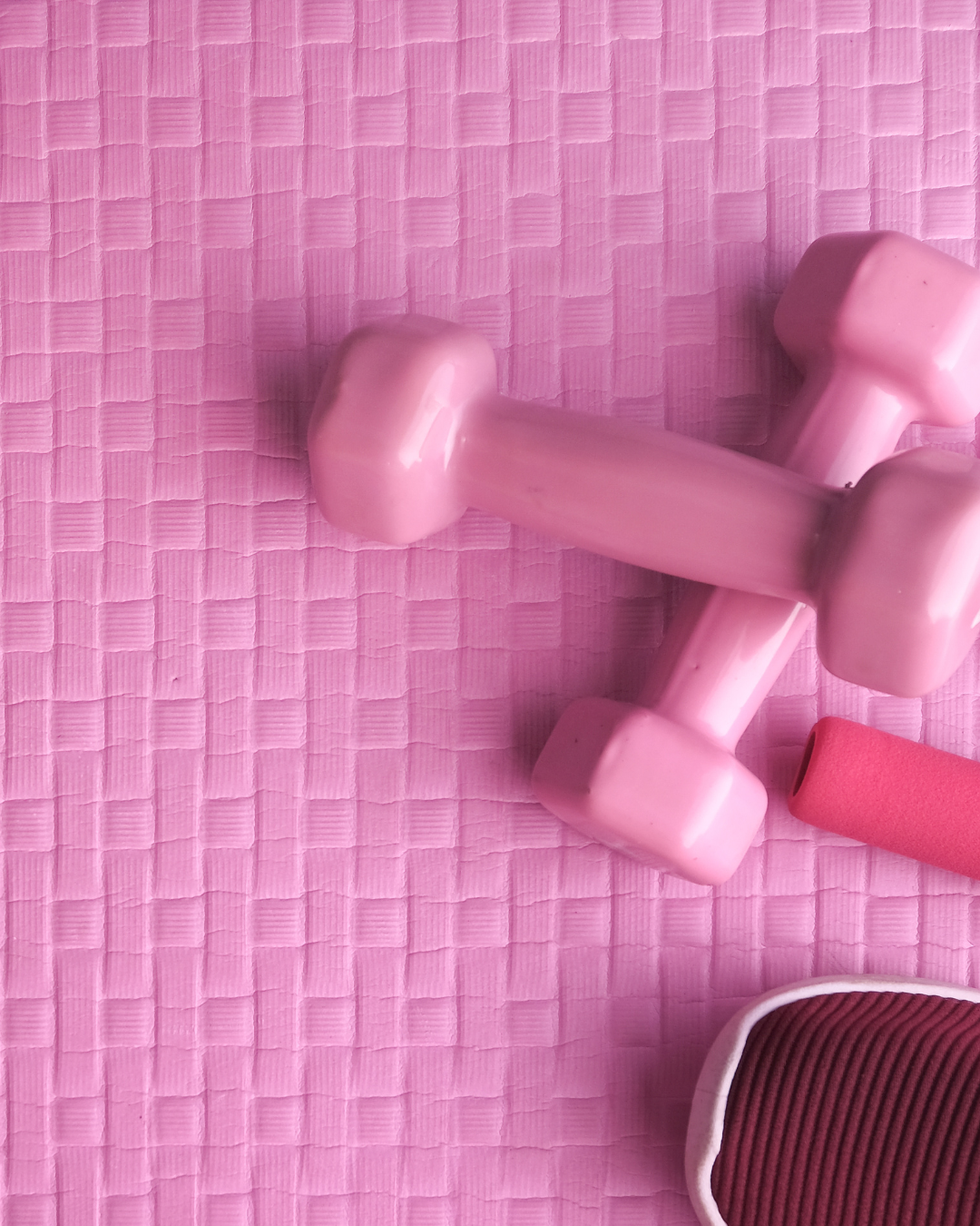 Pink dumbbells on a pink textured yoga mat with a pink foam roller and part of a black and pink exercise shoe.