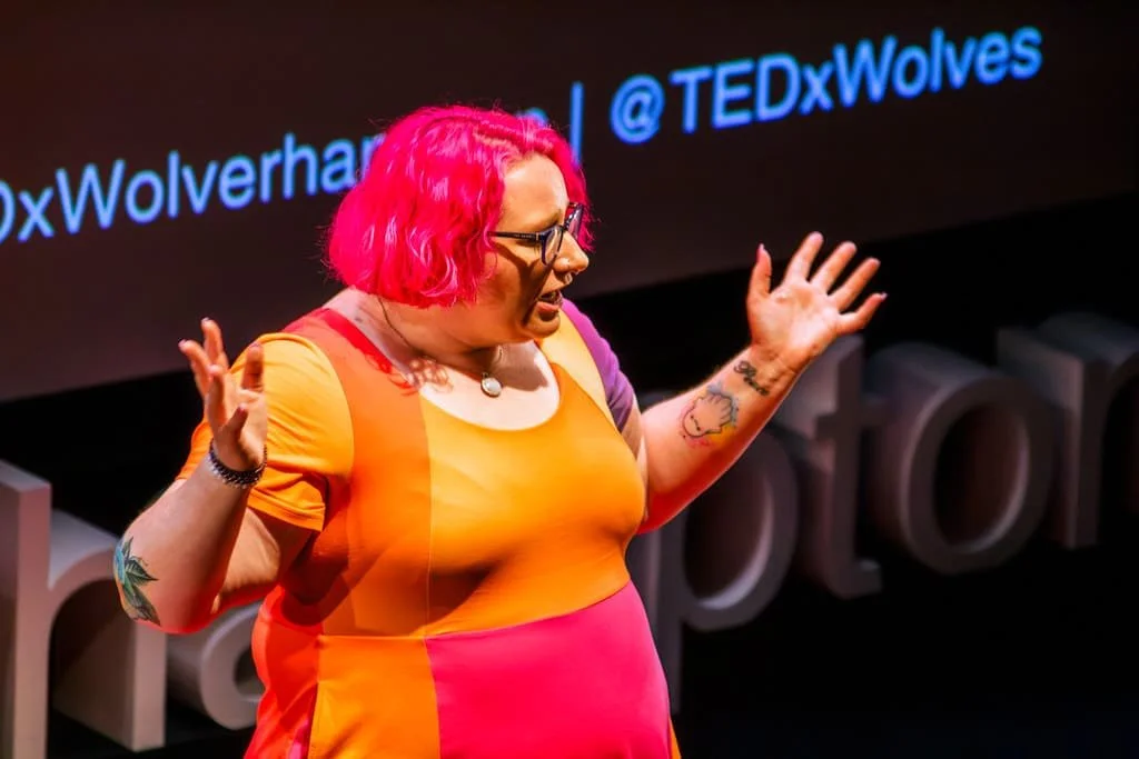 Rhiannon has bright pink hair, wearing glasses and a colourful dress in orange, yellow, and pink, speaking on stage at a TEDx event. She is gesturing with her hands, and a screen with the text '@TEDxWolves' is in the background.