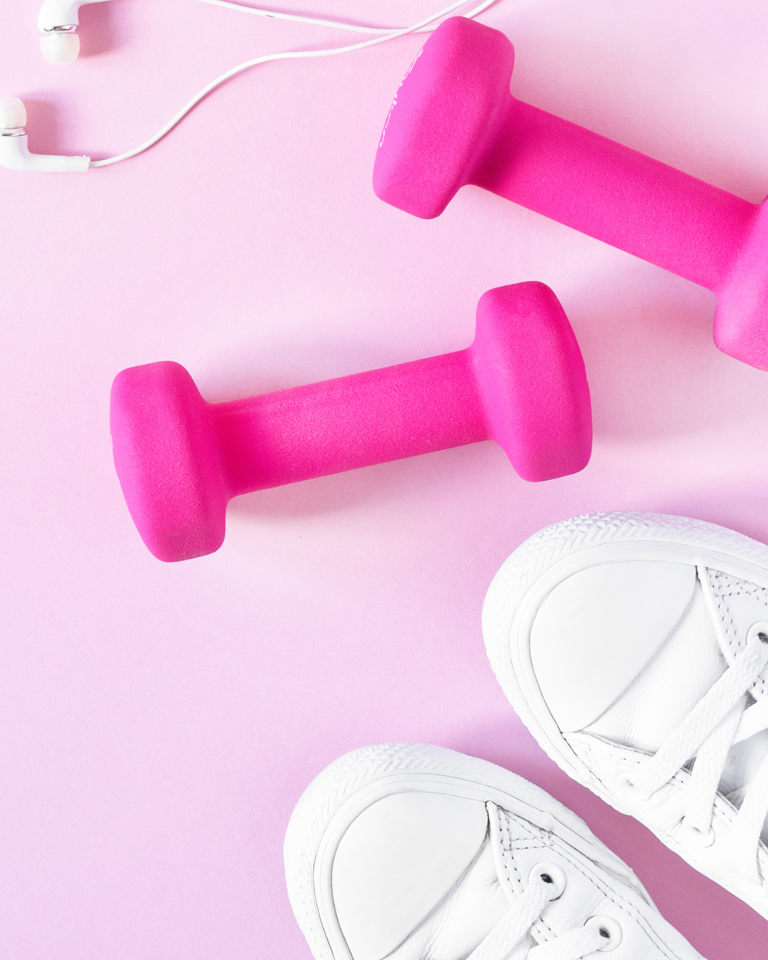 Pink dumbbells, white sneakers, and white earbuds on a pink background.