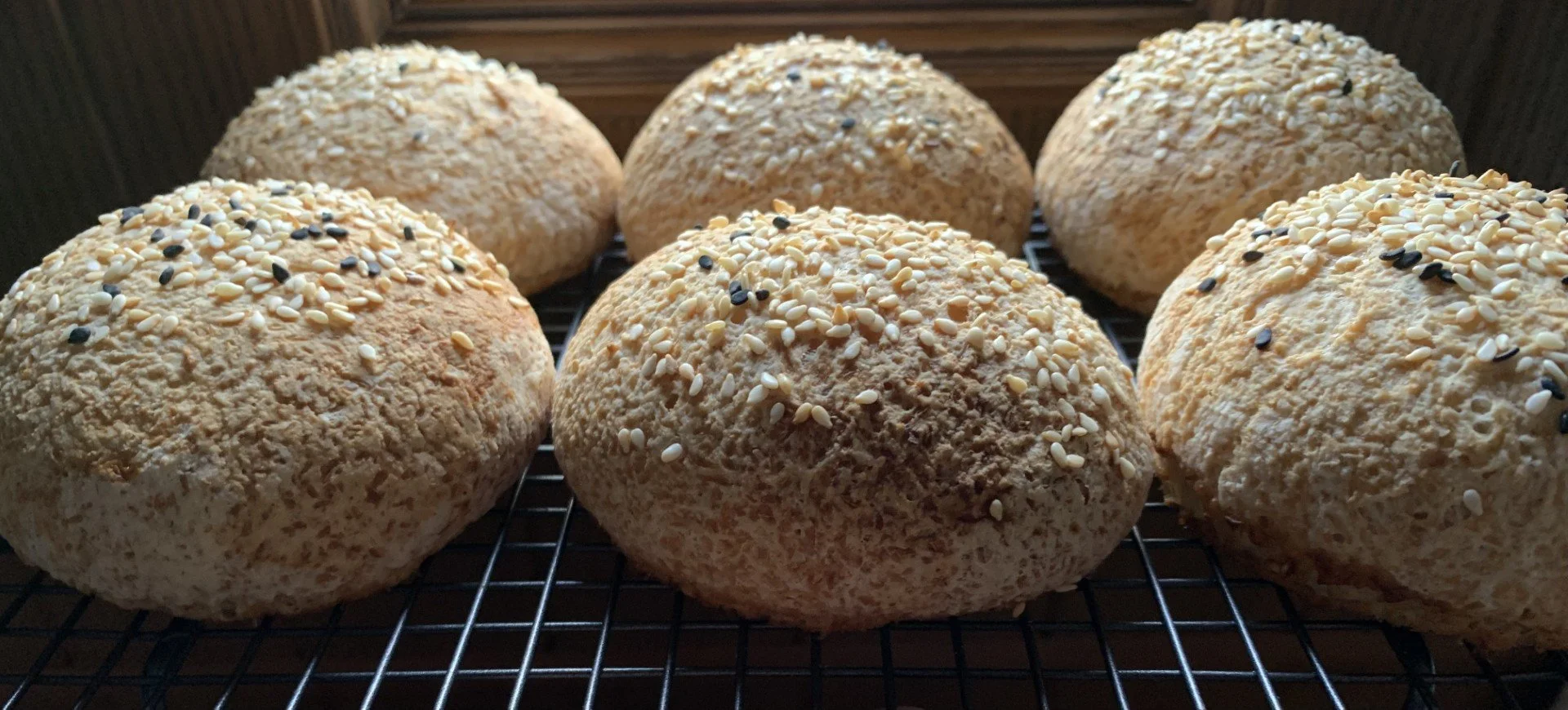 Six gluten-free  buns with sesame seeds cooling on a wire rack.