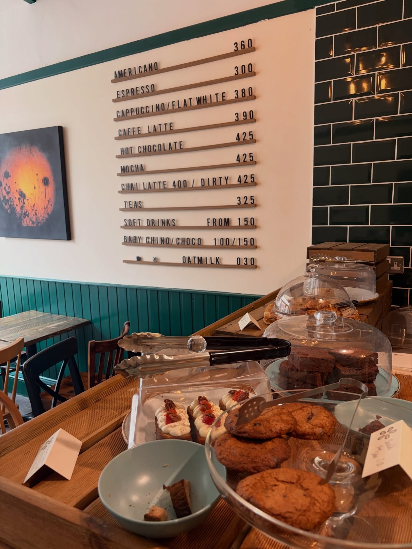 Display of assorted gluten-free  baked goods, gluten-free cookies, and gluten-free desserts on a wooden table in a cafe, with a menu board in the background listing coffee and tea options.