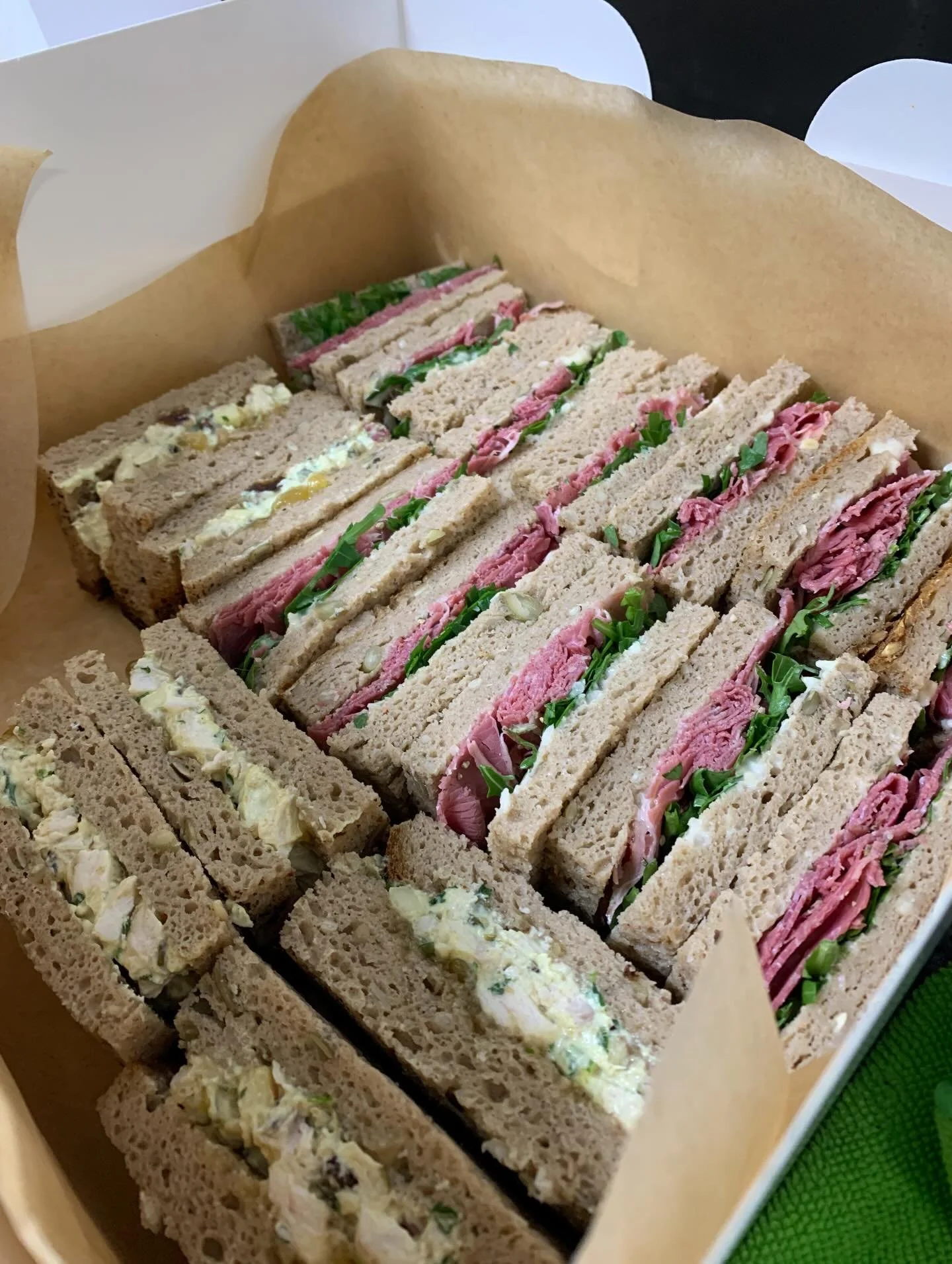 A box of assorted gluten-free  sandwiches made with whole wheat bread, filled with meats, greens, and various spreads.