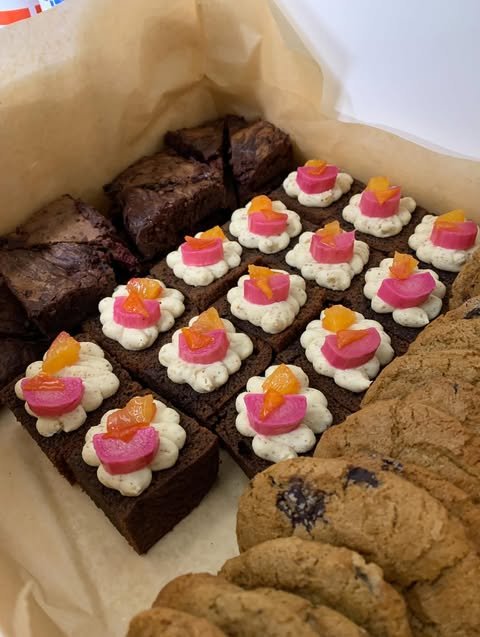 Assorted gluten-free cookies and gluten-free brownies in a box. The box contains chocolate brownies topped with colorful frosting and candy pieces, and oatmeal cookies with chocolate chips.