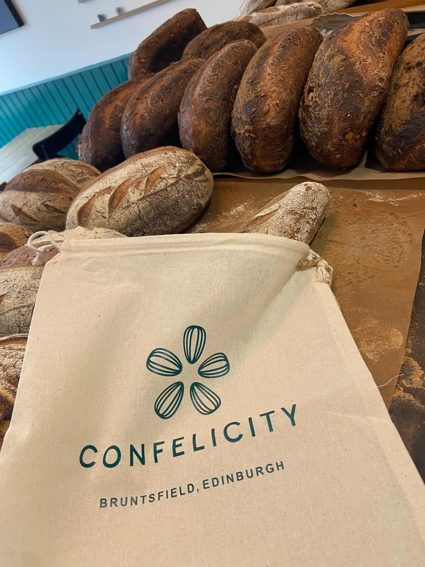 Display of various freshly baked gluten-free bread loaves on a table at Conelficty bakery in Bruntsfield, Edinburgh, with a branded cloth bag in the foreground.