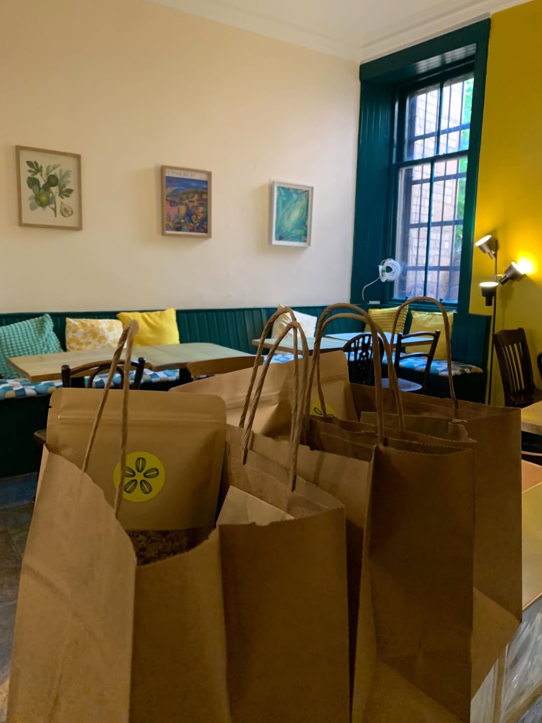 Brown paper bags with yellow stickers on a table inside a cafe or restaurant, with colorful pillows, paintings, a large window, and a floor lamp in the background.