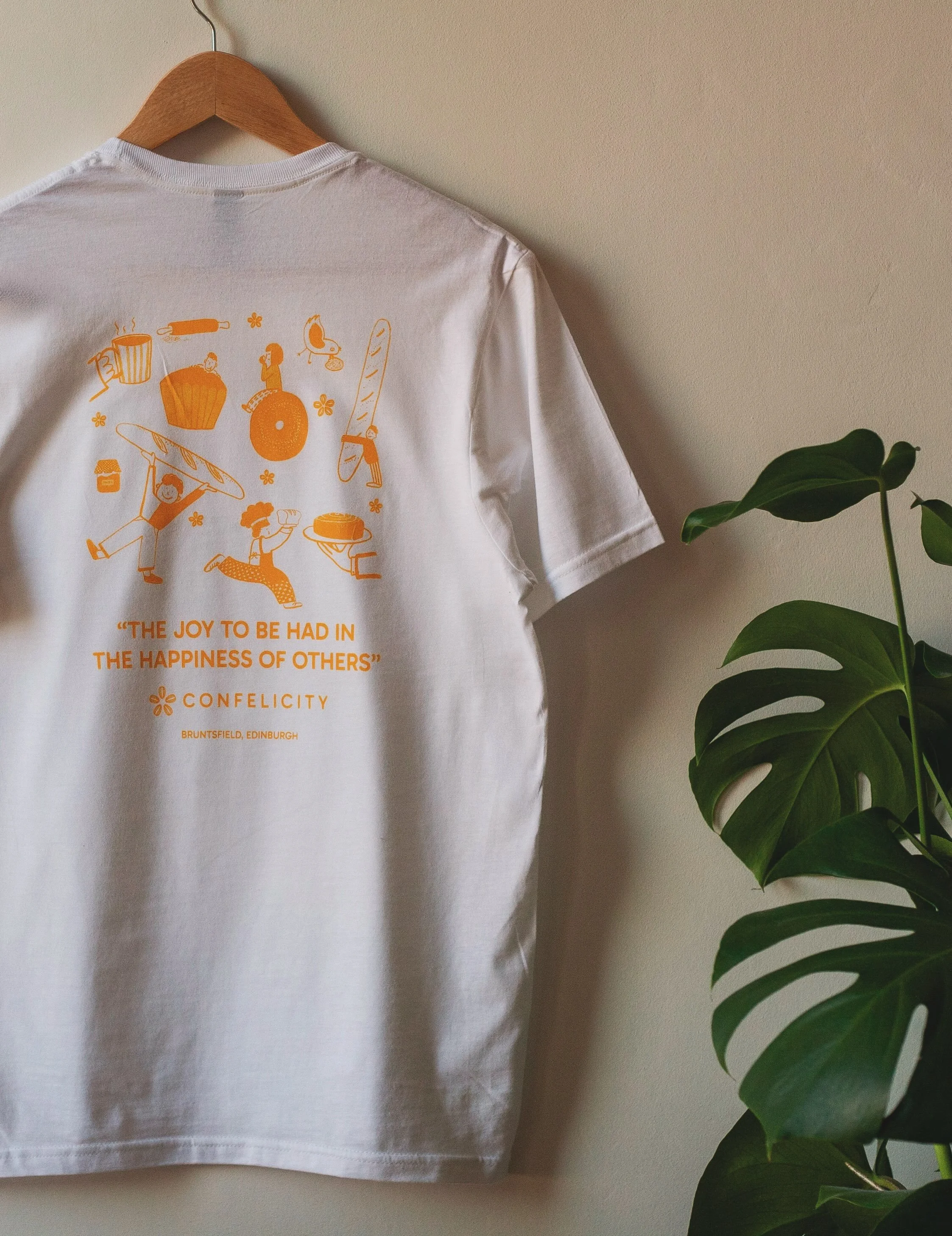 White t-shirt hanging on a wooden hanger next to a green leafy plant, with orange graphic design and text on the shirt.
