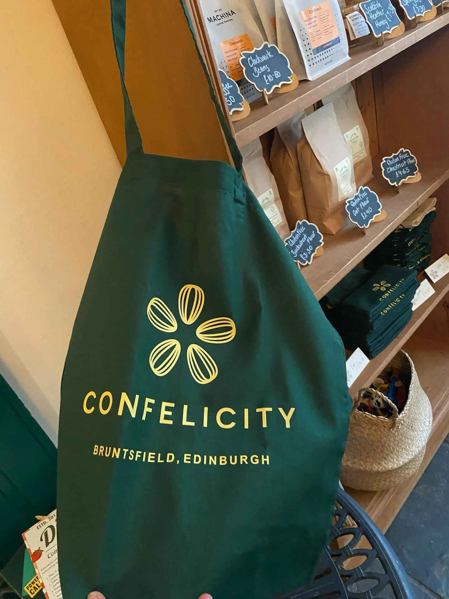 A retail display shelf with packaged food items and handwritten price tags, and a green tote bag with the branding of Confecticty, located in Edinburgh.